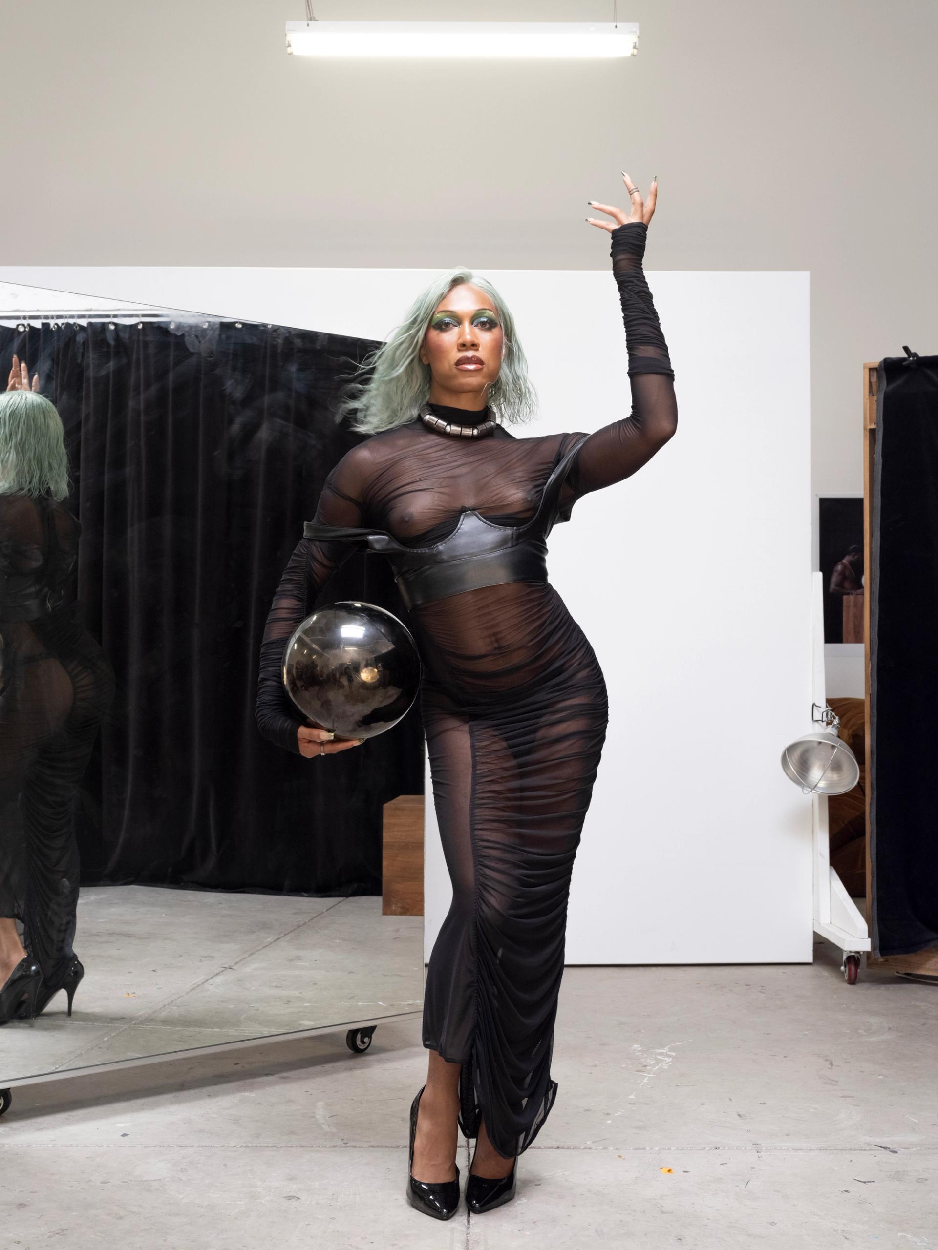 Person with mint-green hair poses in a sheer black dress, holding a reflective sphere in a studio.