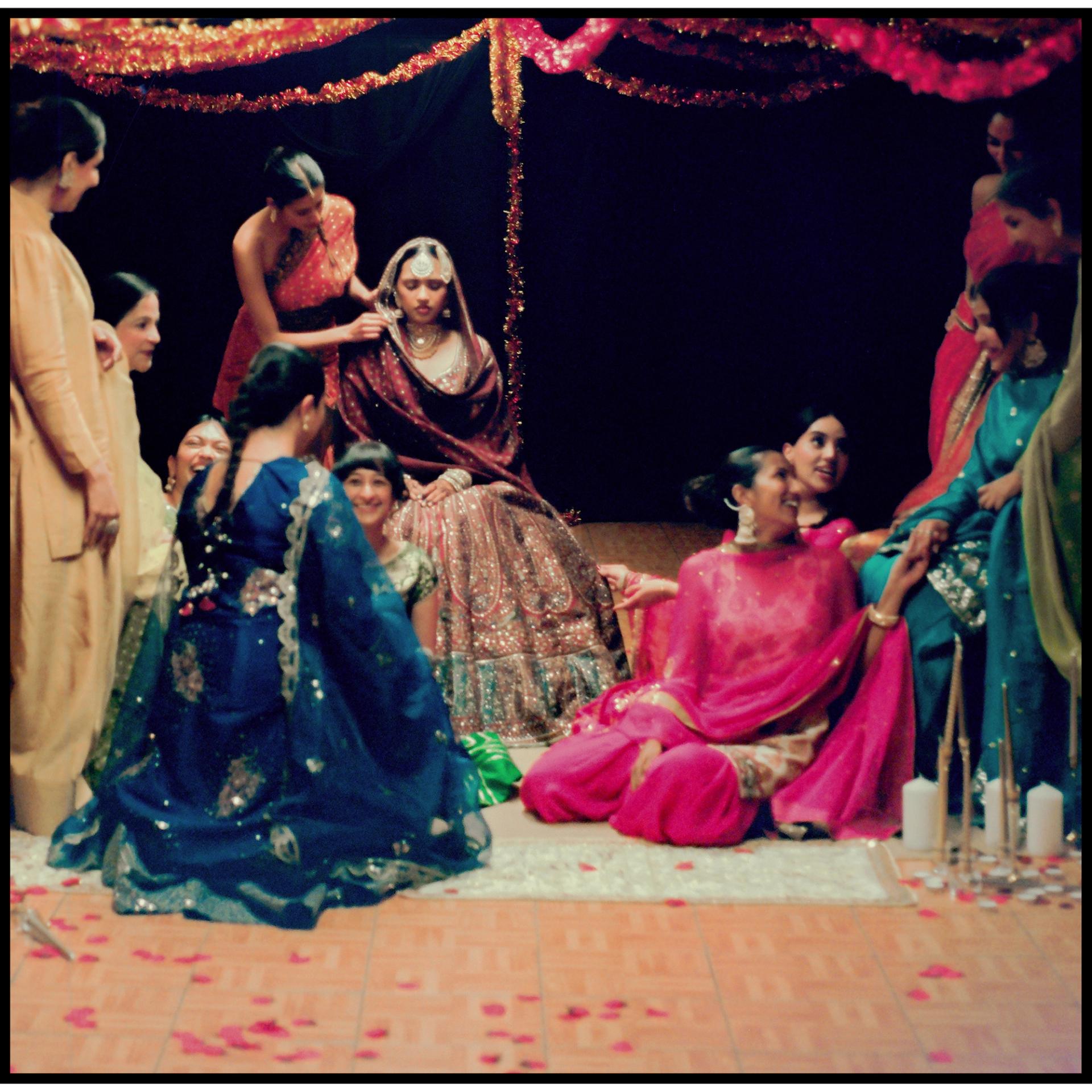 A group of performers in colorful South Asian attire gather around a central bridal figure seated beneath tinsel garlands, with rose petals and candles on the floor around them