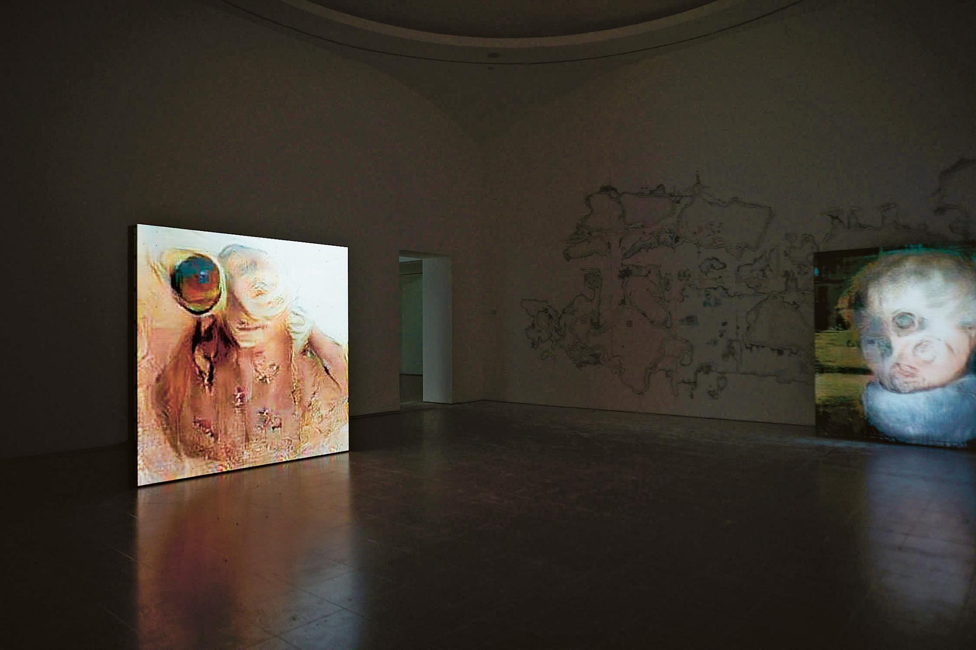 A darkened circular gallery space with two large projected video works showing distorted figurative imagery, and an abstract line drawing on the wall between them