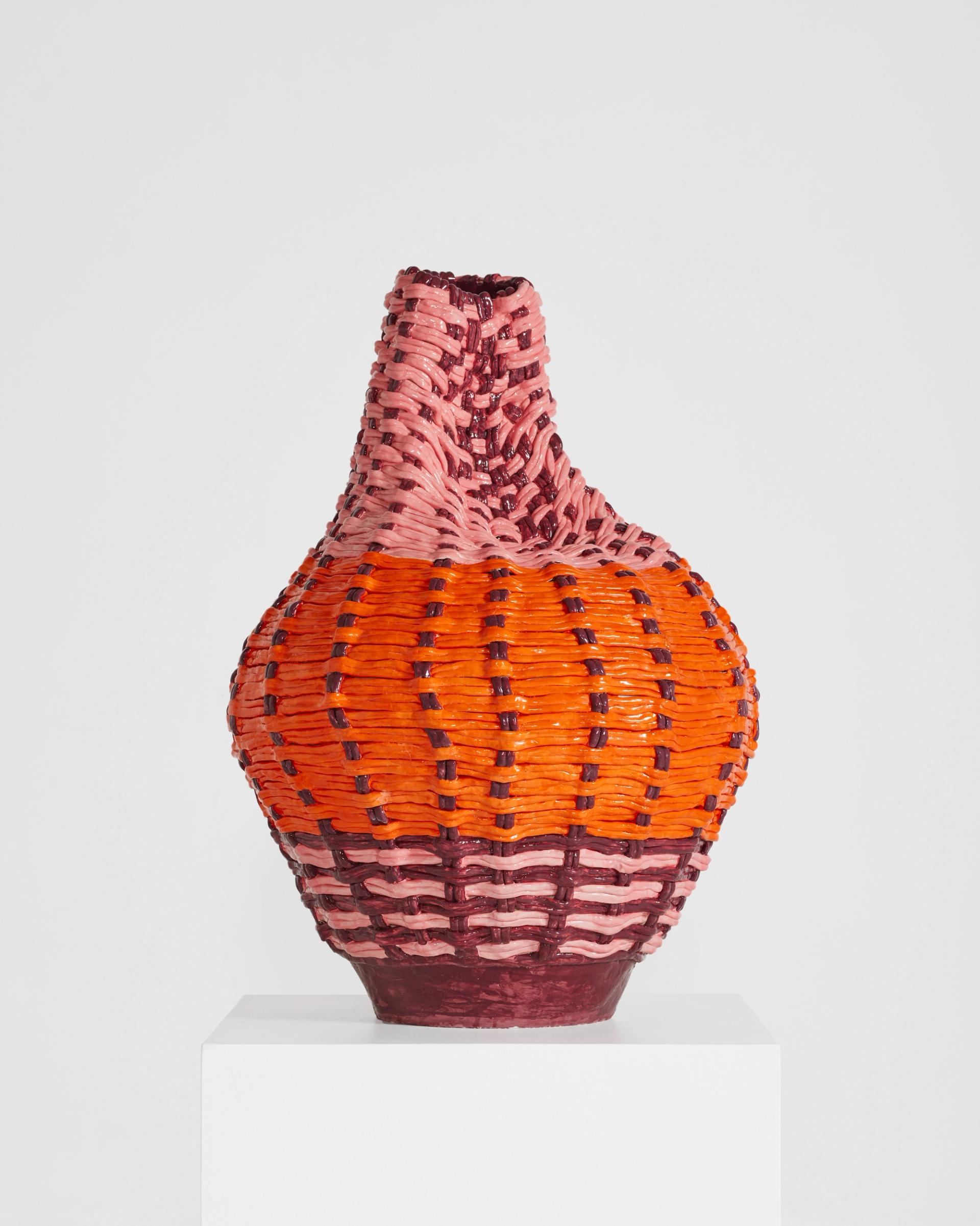 An orange, pink, and maroon ceramic vessel with a woven texture sits on a white pedestal in front of a white background.