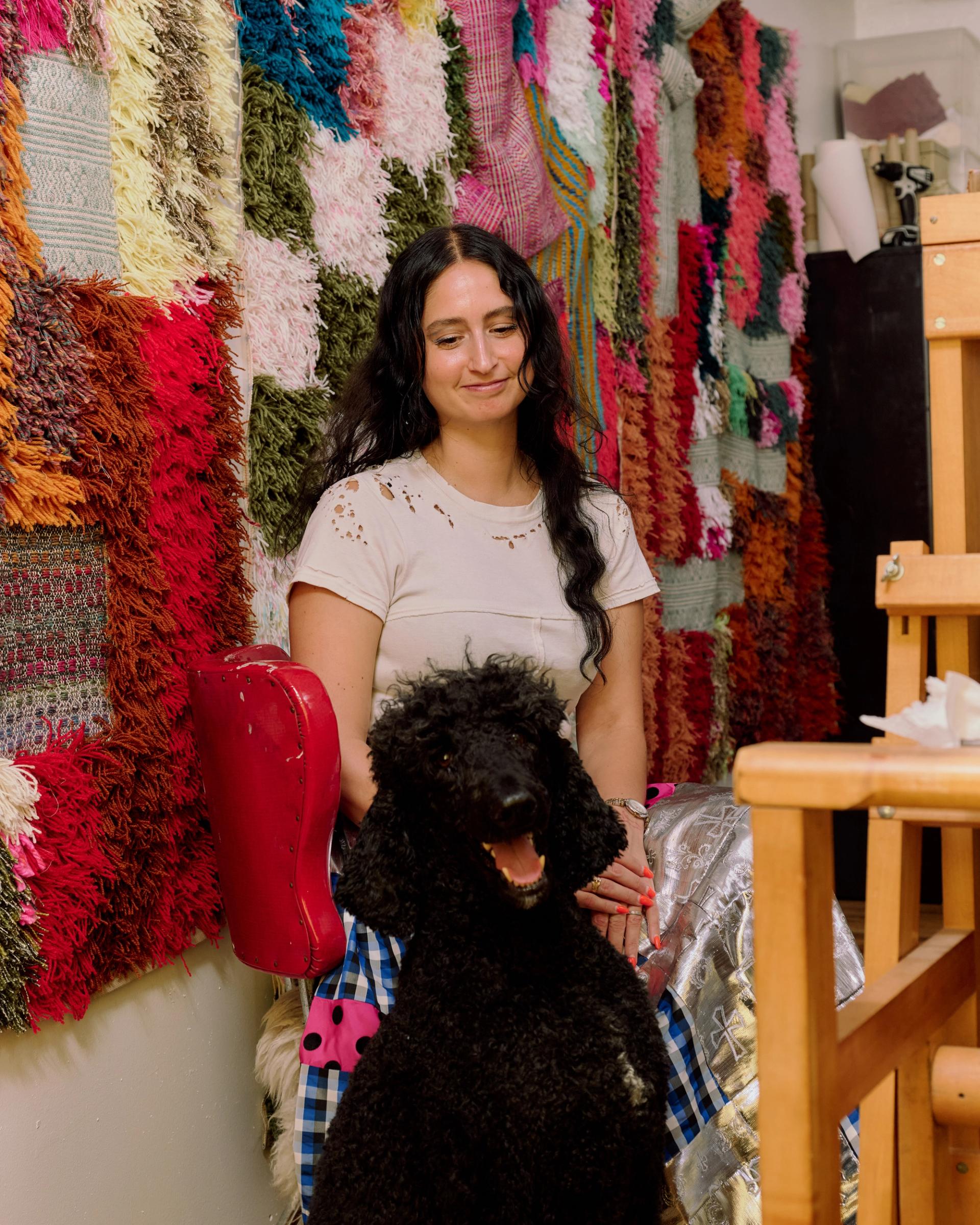A smiling woman seated behind a large black curly-haired dog in a studio hung with vibrant multicolored textile artworks