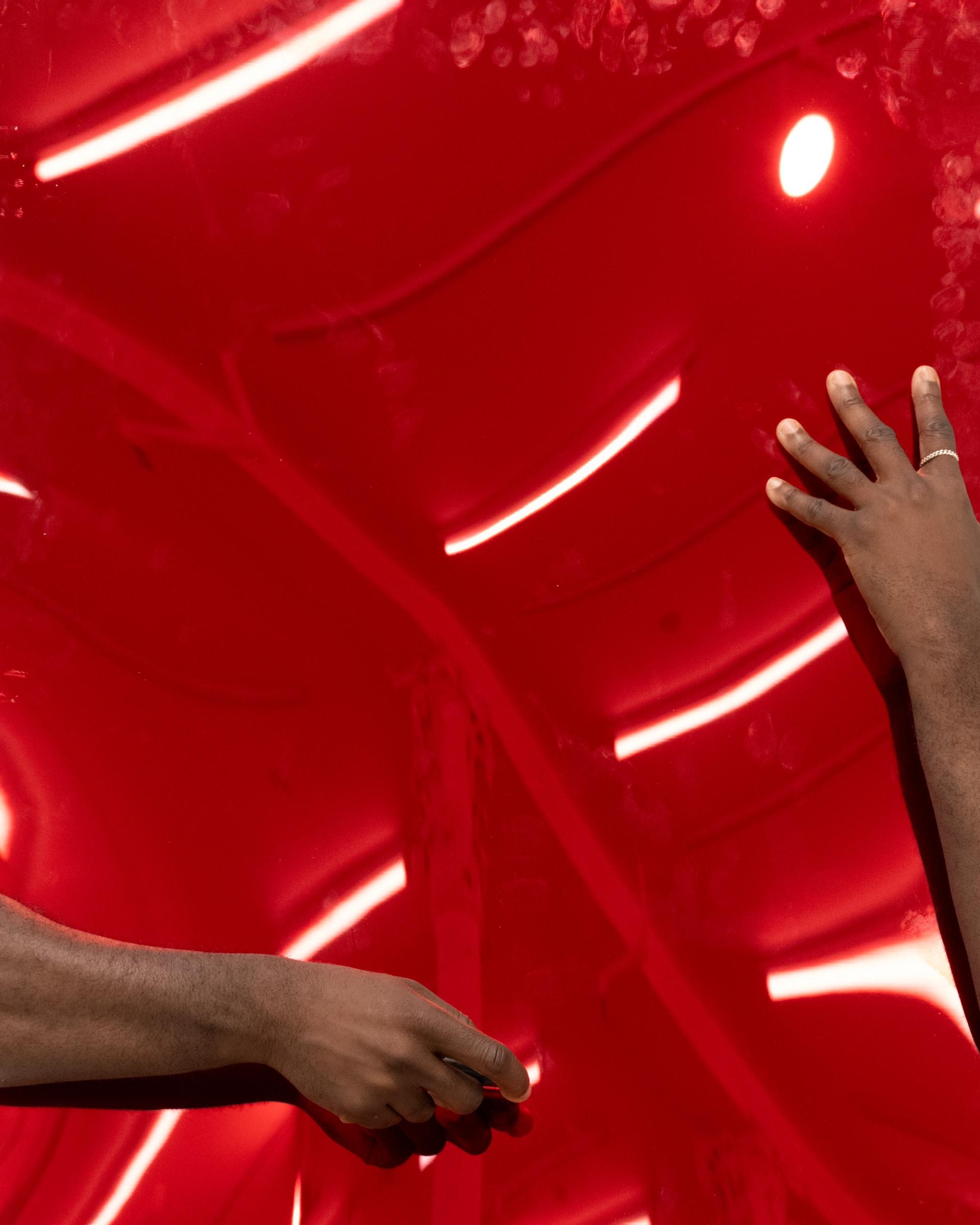 Two hands reach across a glossy red reflective surface marked with fingerprints and light streaks.