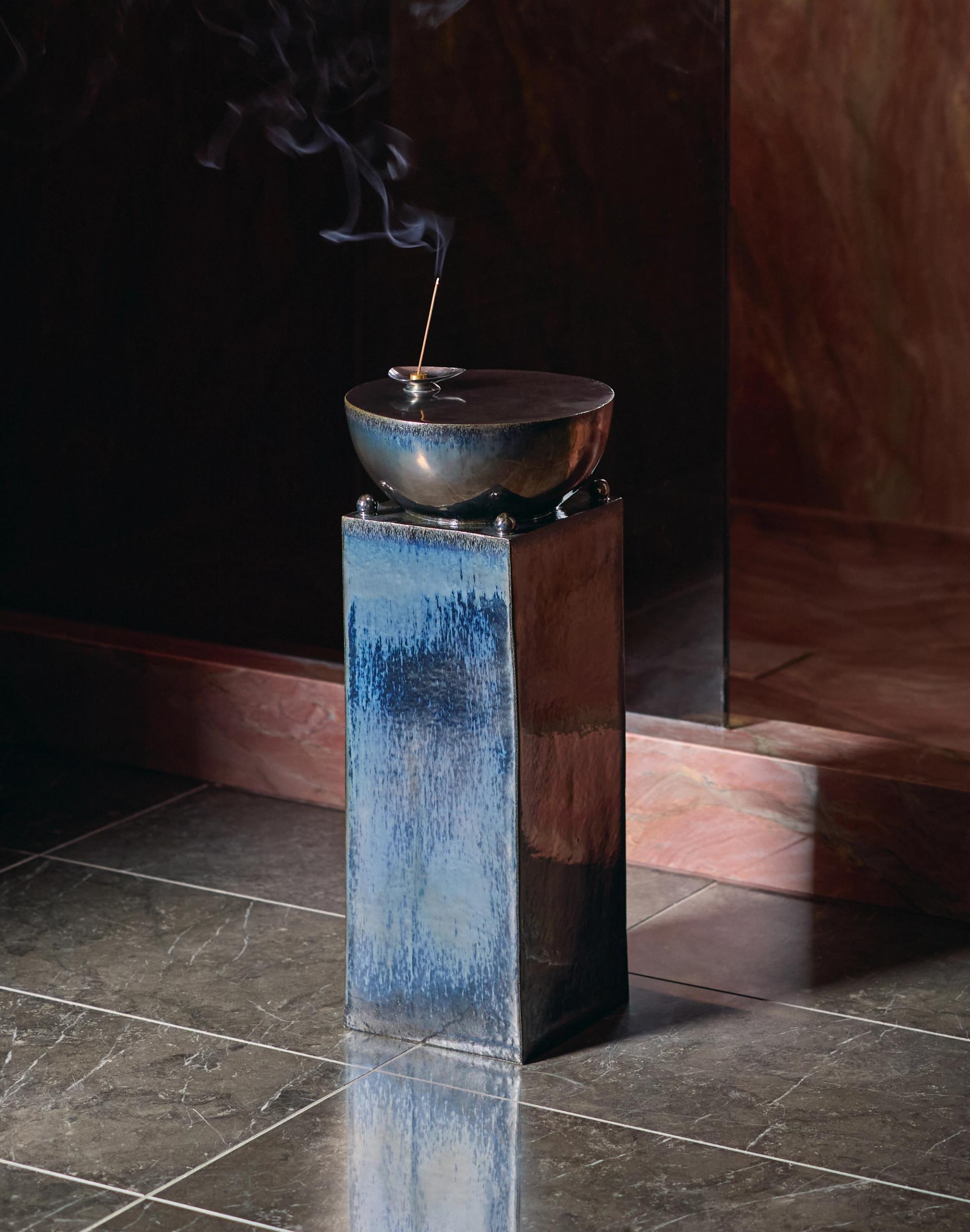 Smoking incense vessel resting on tall pedestal in dimly lit interior with reflective stone flooring.