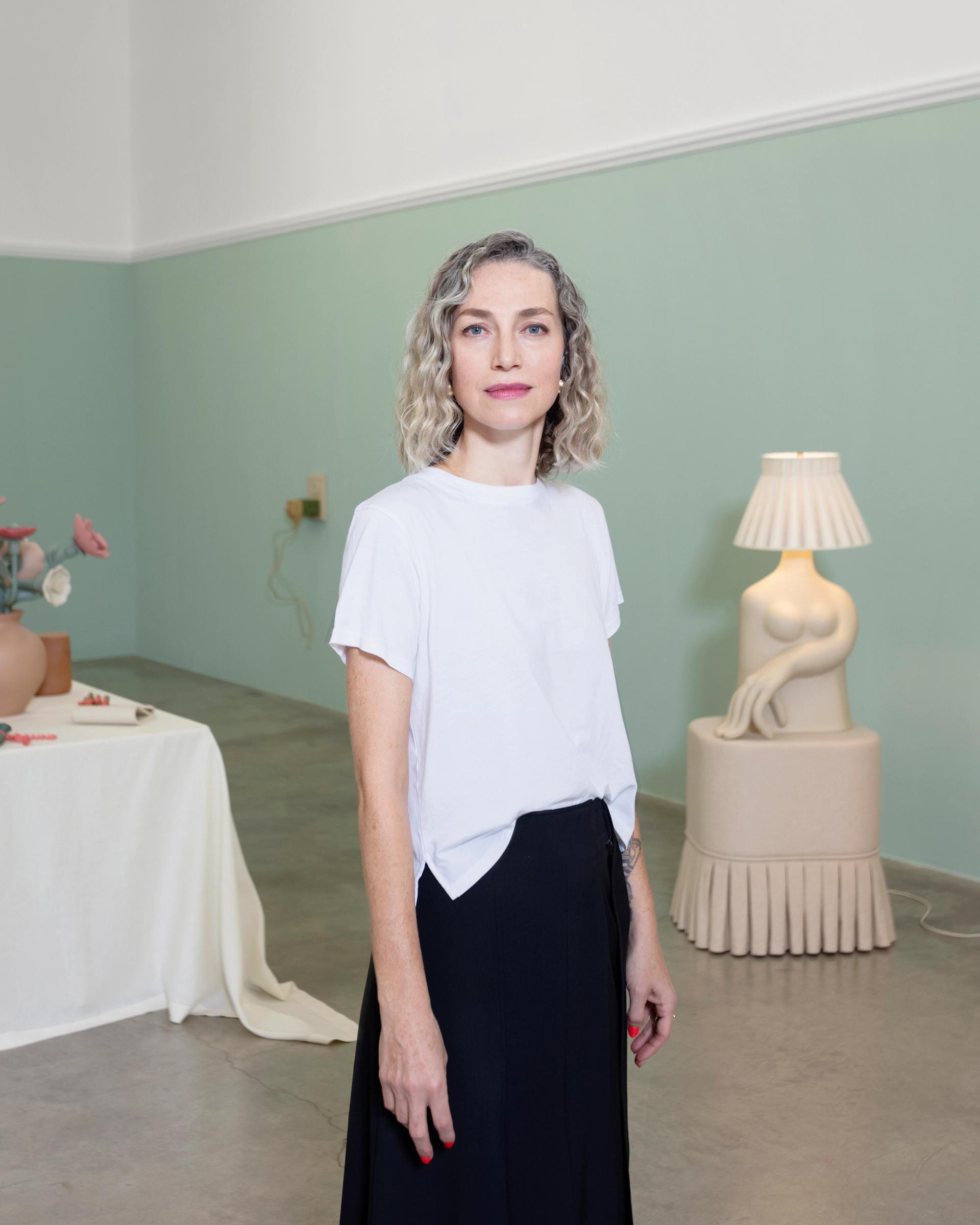 A woman with curly silver-blond hair in a white T-shirt and black skirt stands in a gallery space, with a ceramic figurative lamp sculpture and a draped table visible behind her.