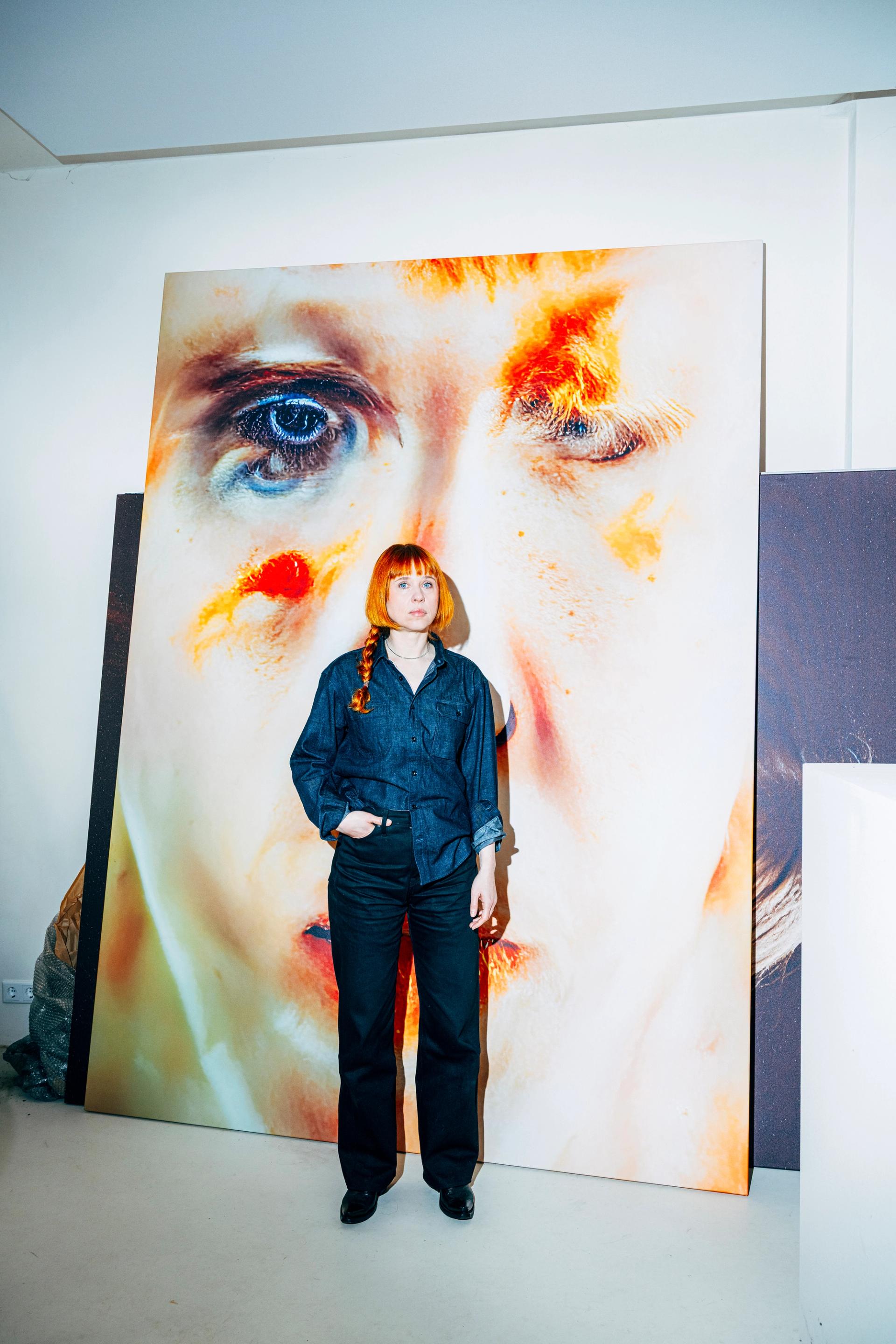 A woman with a red braid wearing a dark denim outfit stands in an art studio in front of a large-scale painting depicting an extreme close-up of a face with vivid orange and red tones.