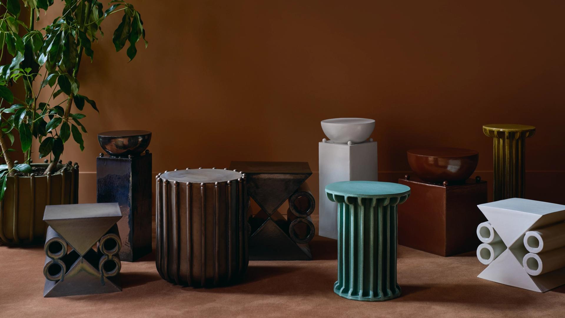 Arrangement of colorful ceramic columns and stools against an orange wall with a potted plant in one corner.