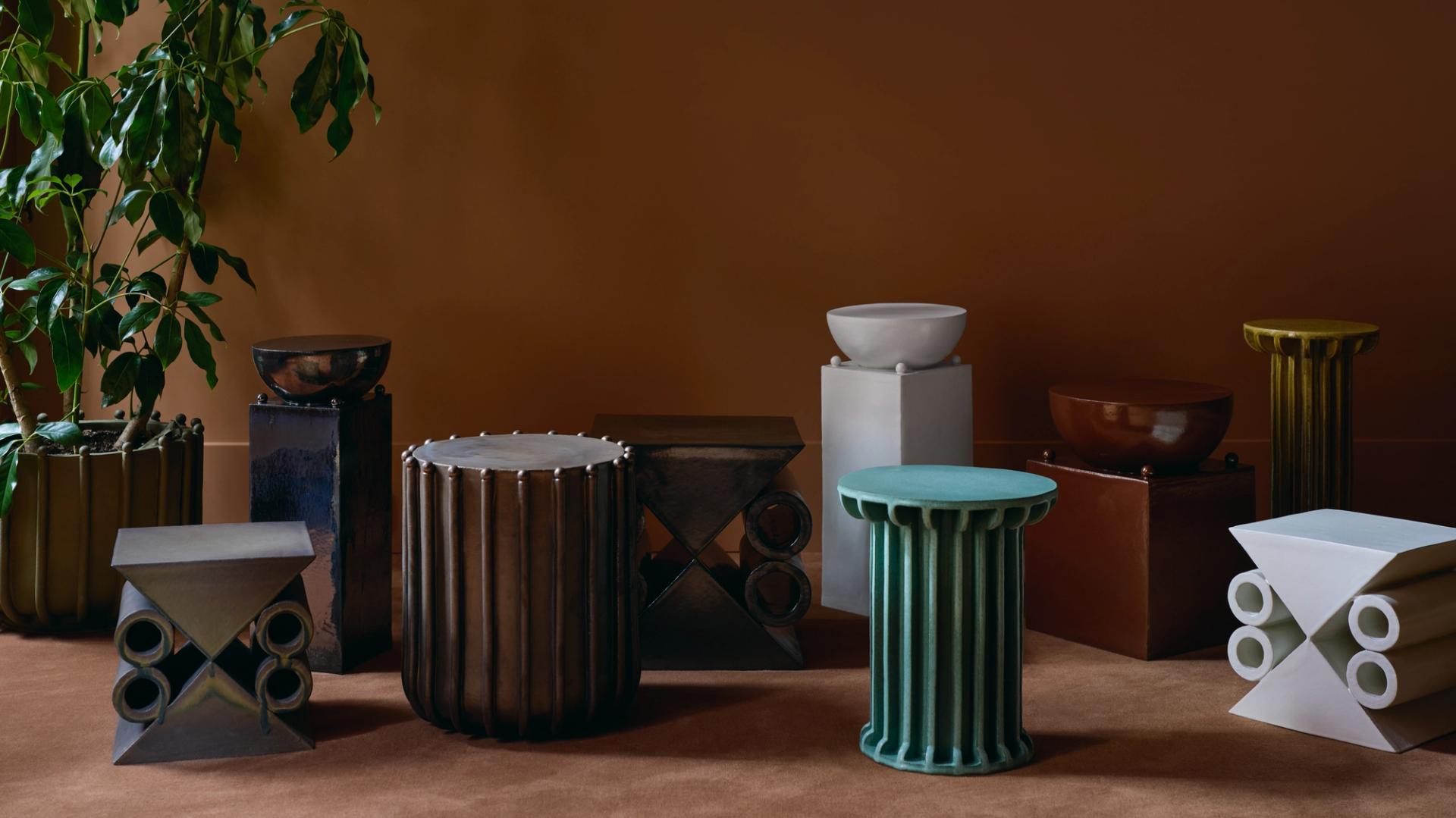 Arrangement of colorful ceramic columns and stools against an orange wall with a potted plant in one corner.