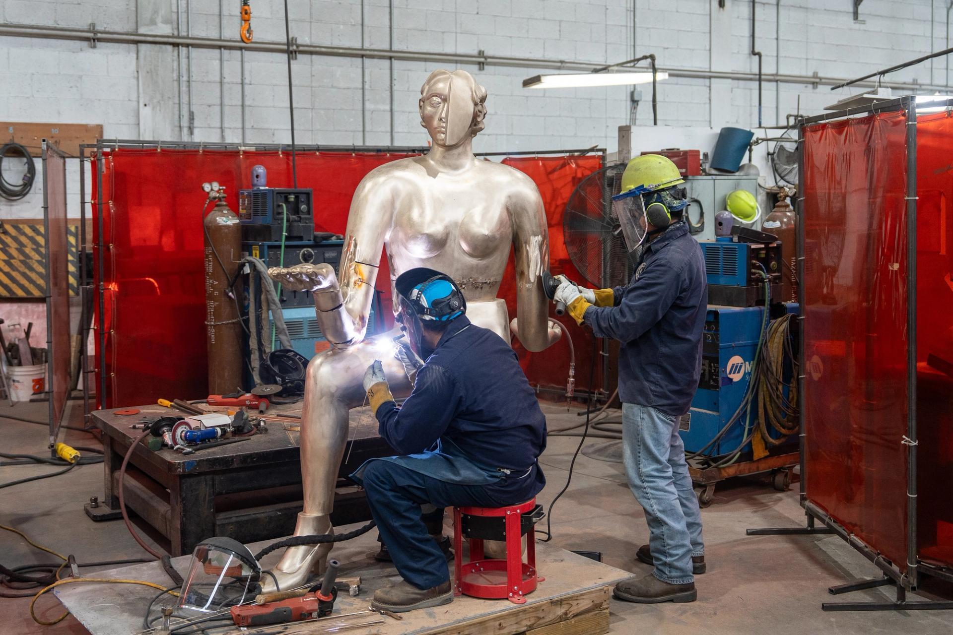 Two workers in protective gear weld and finish a large polished metal figurative sculpture of a seated woman in an industrial fabrication workshop