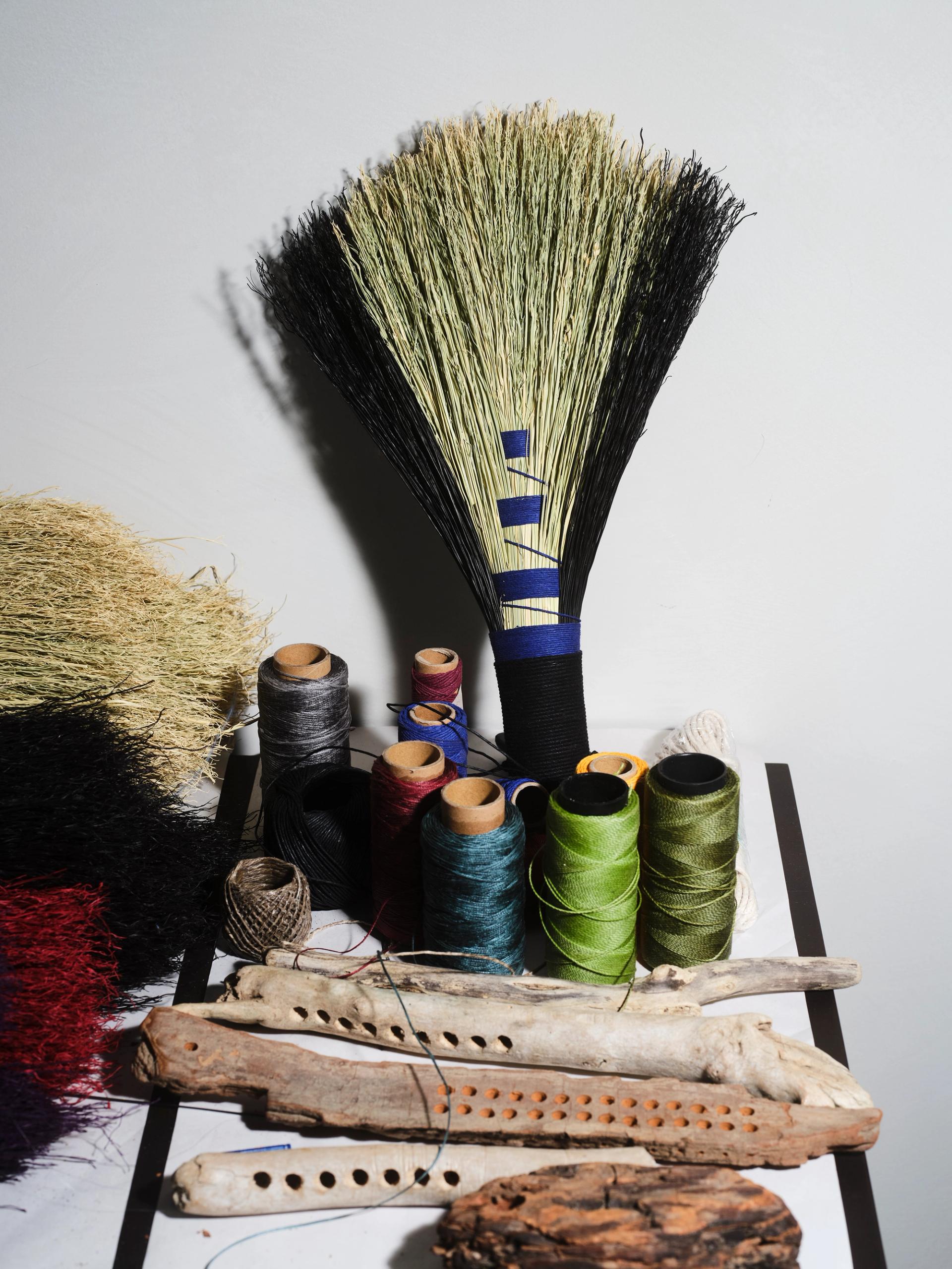 Finished broom with black and natural fibers displayed with colorful thread spools and wooden tools on a table.