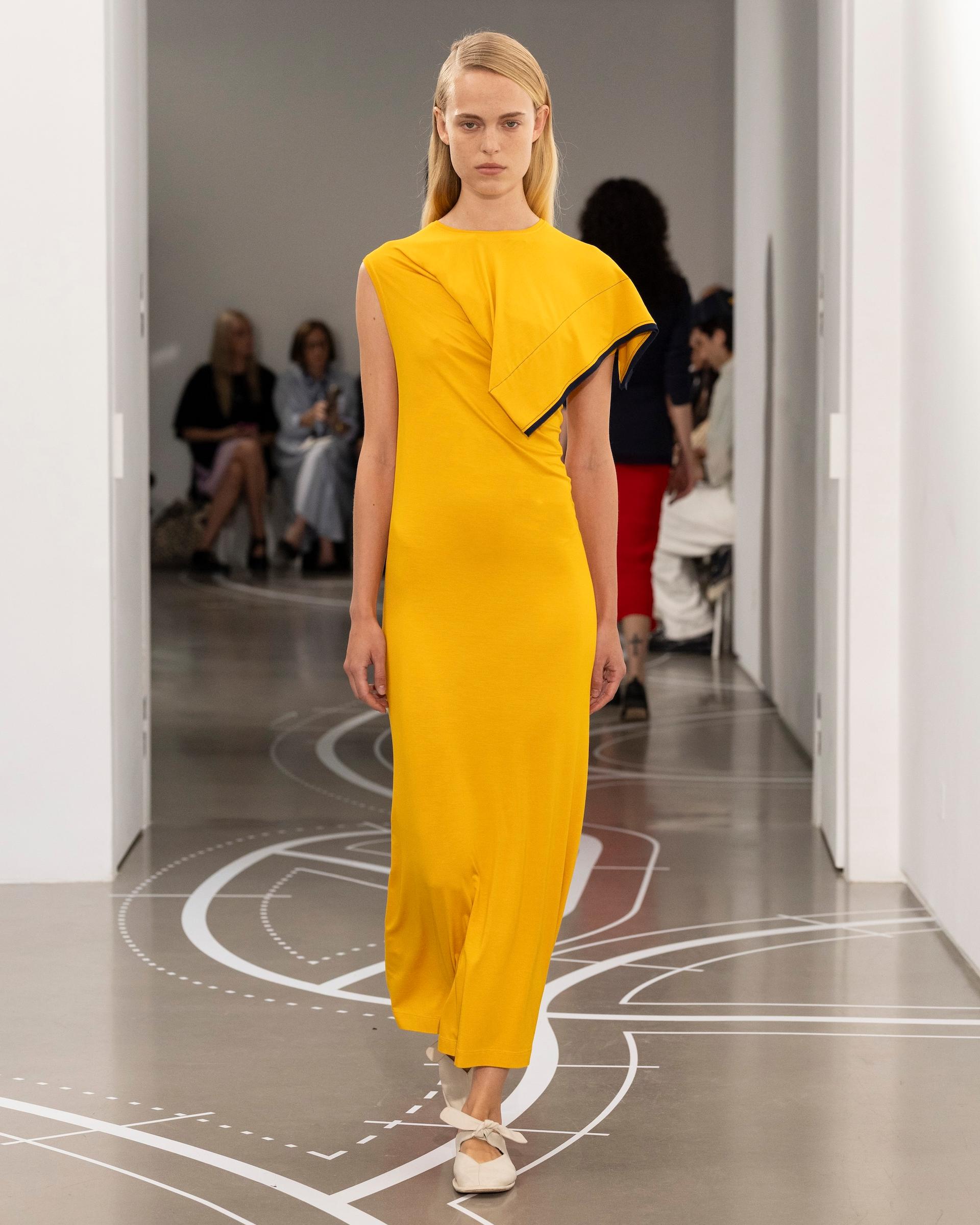 A model walks a runway in a mustard yellow maxi dress with an asymmetric folded panel detail and navy trim, paired with white bow-front flats.