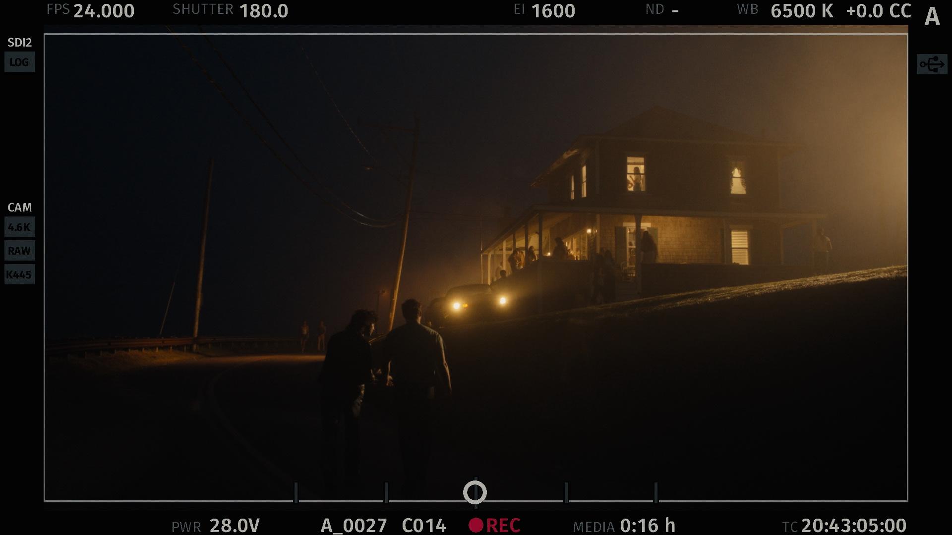 Behind-the-scenes still from a camera monitor showing two silhouetted figures walking a dark road toward a glowing, fog-lit house at night, with a truck’s headlights on and people gathered on the porch.