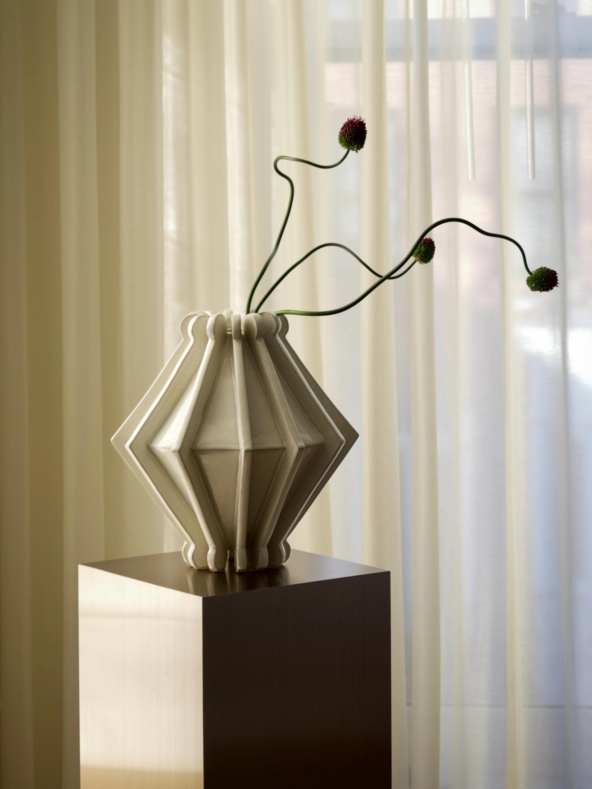 Geometric ceramic vase with long-stemmed flowers displayed on pedestal before sheer curtains.