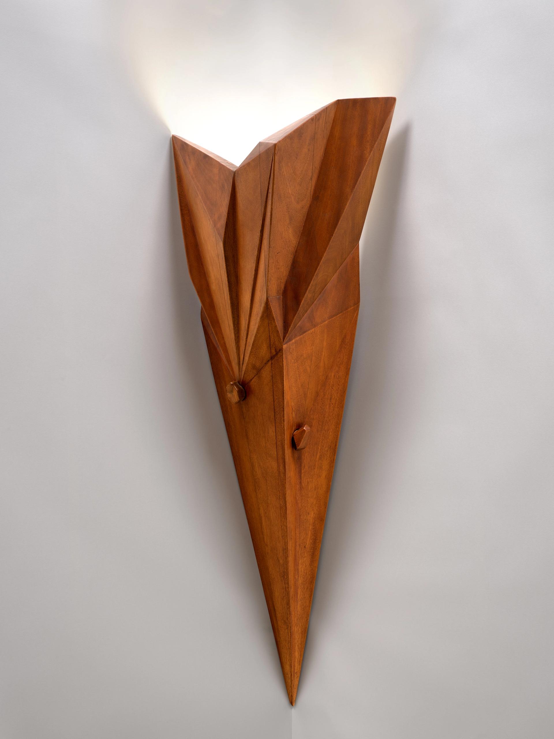 Geometric carved wooden wall sconce with faceted, angular planes tapering to a point, mounted on a white wall and casting soft upward light.