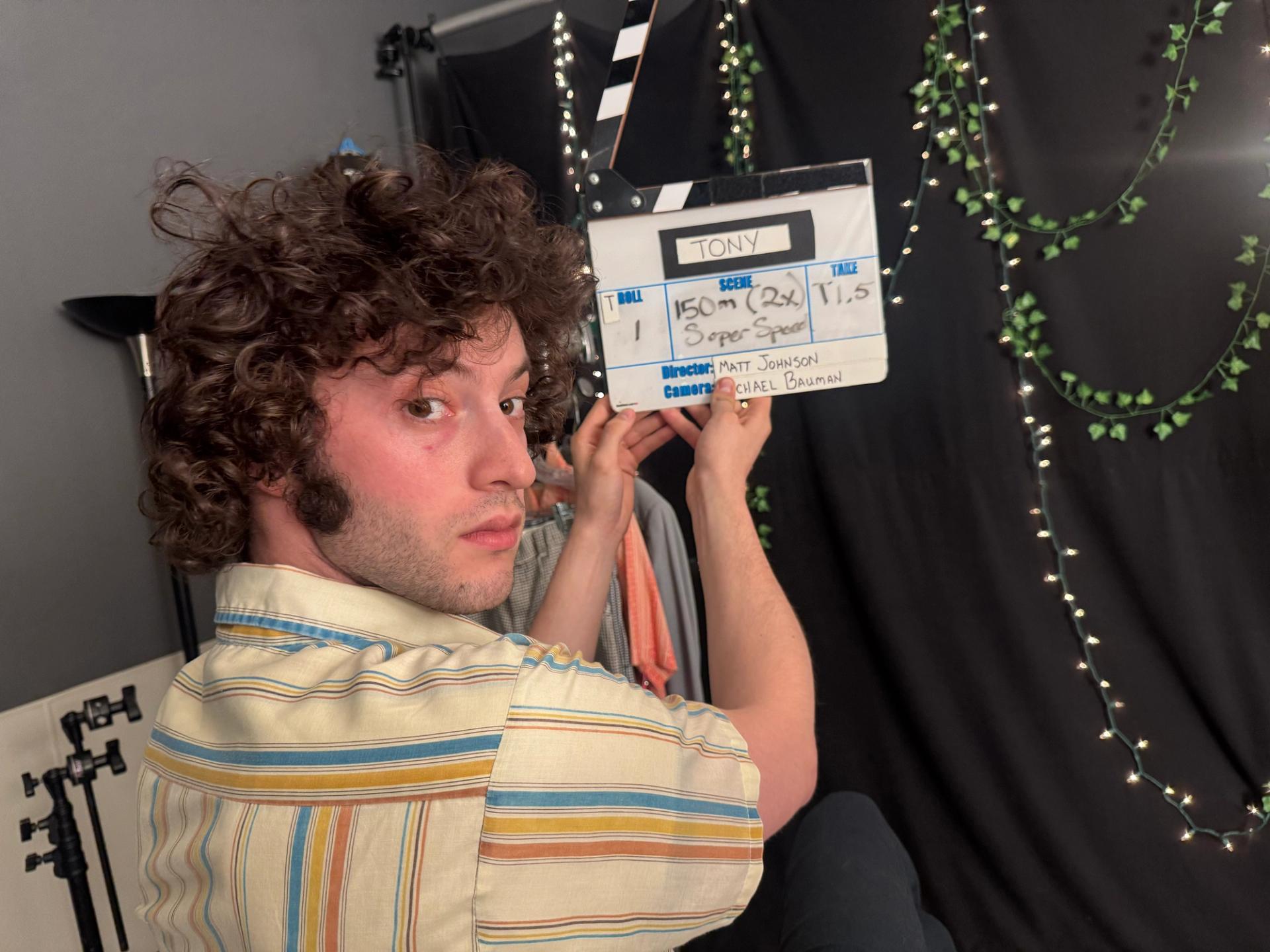 A curly-haired man in a striped shirt looking back over his shoulder while holding up a film clapperboard reading “Tony,” with scene and director information