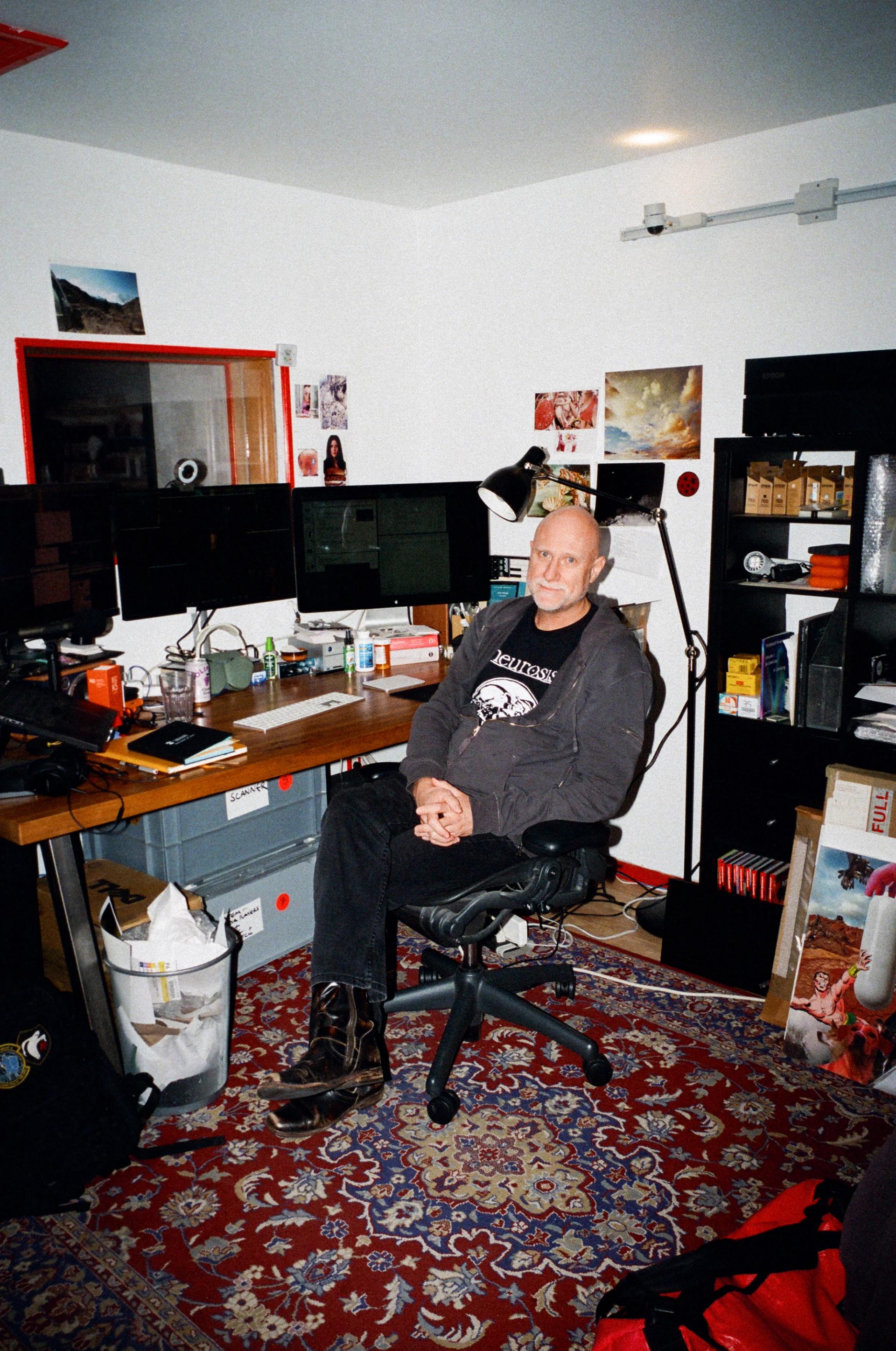 A bald man sits in a desk chair in front of a desk with three computer monitors.