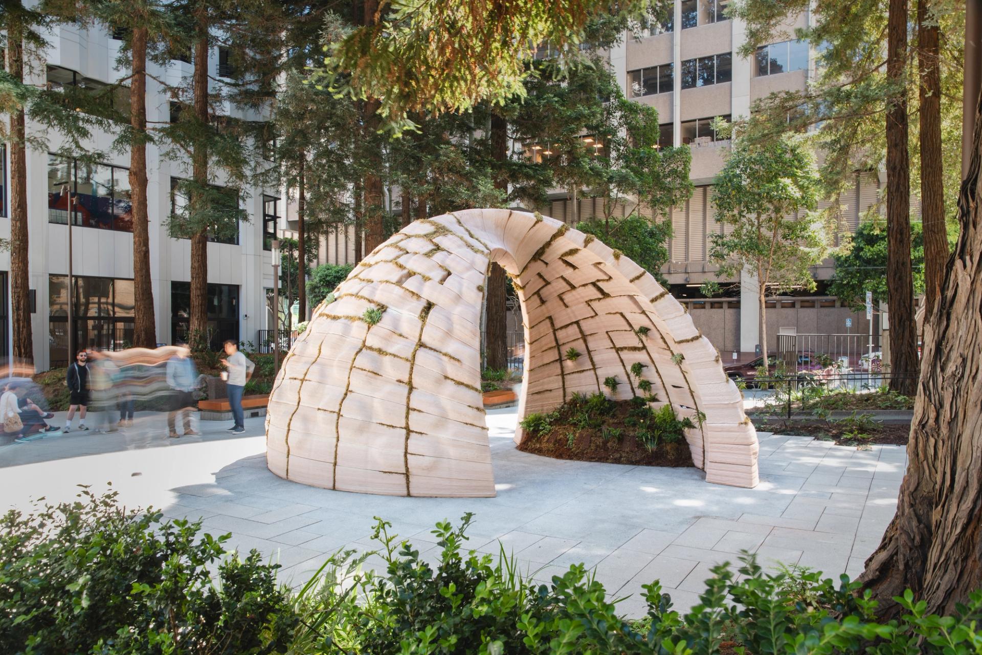 A large arch-shaped structure of pale stacked blocks with moss and plants growing in its joints, installed in an urban plaza among tall redwood trees with passersby visible nearby
