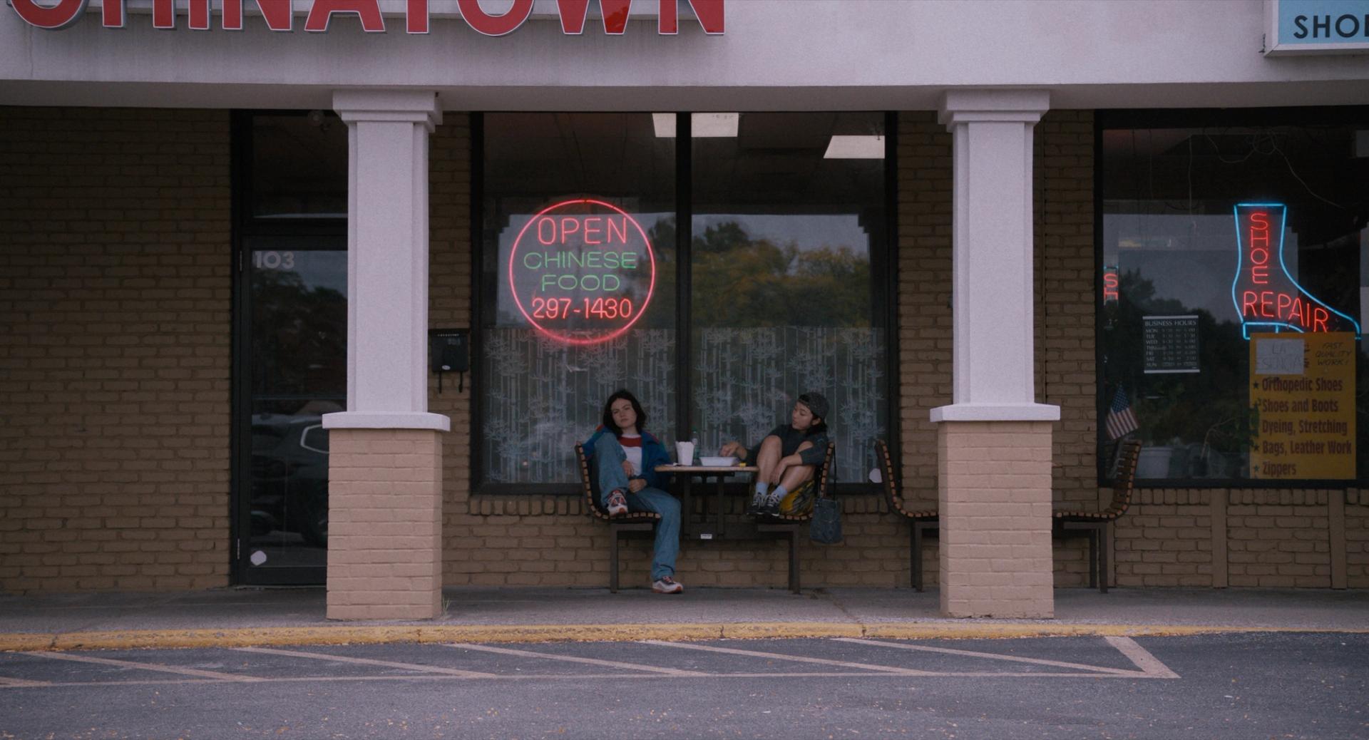Two people sit outside a strip-mall storefront beneath a glowing Chinese-food neon sign.