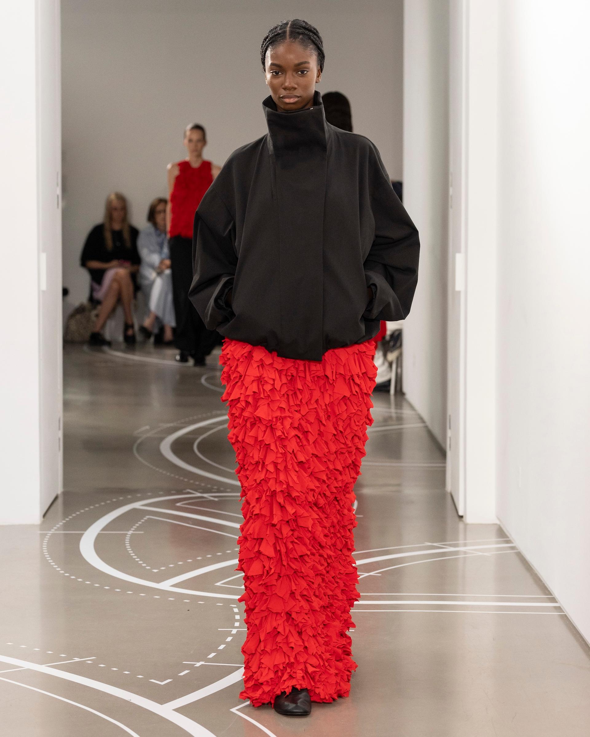 A model walks a runway wearing an oversized black turtleneck jacket paired with a floor-length red heavily ruffled skirt, with seated guests visible in the background
