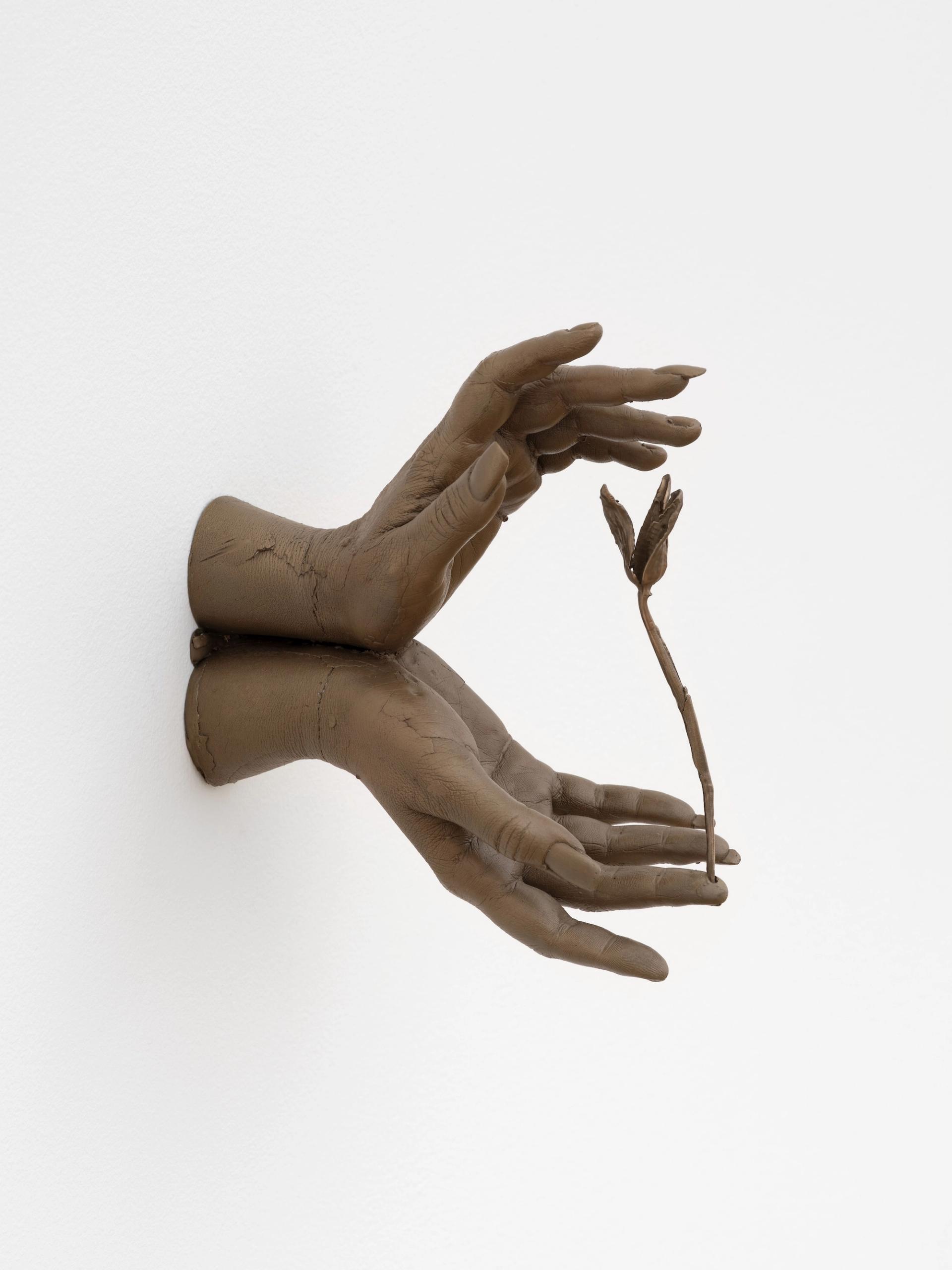 A bronze wall-mounted sculpture of two cast hands emerging from a white wall, cradling a small flower stem between them