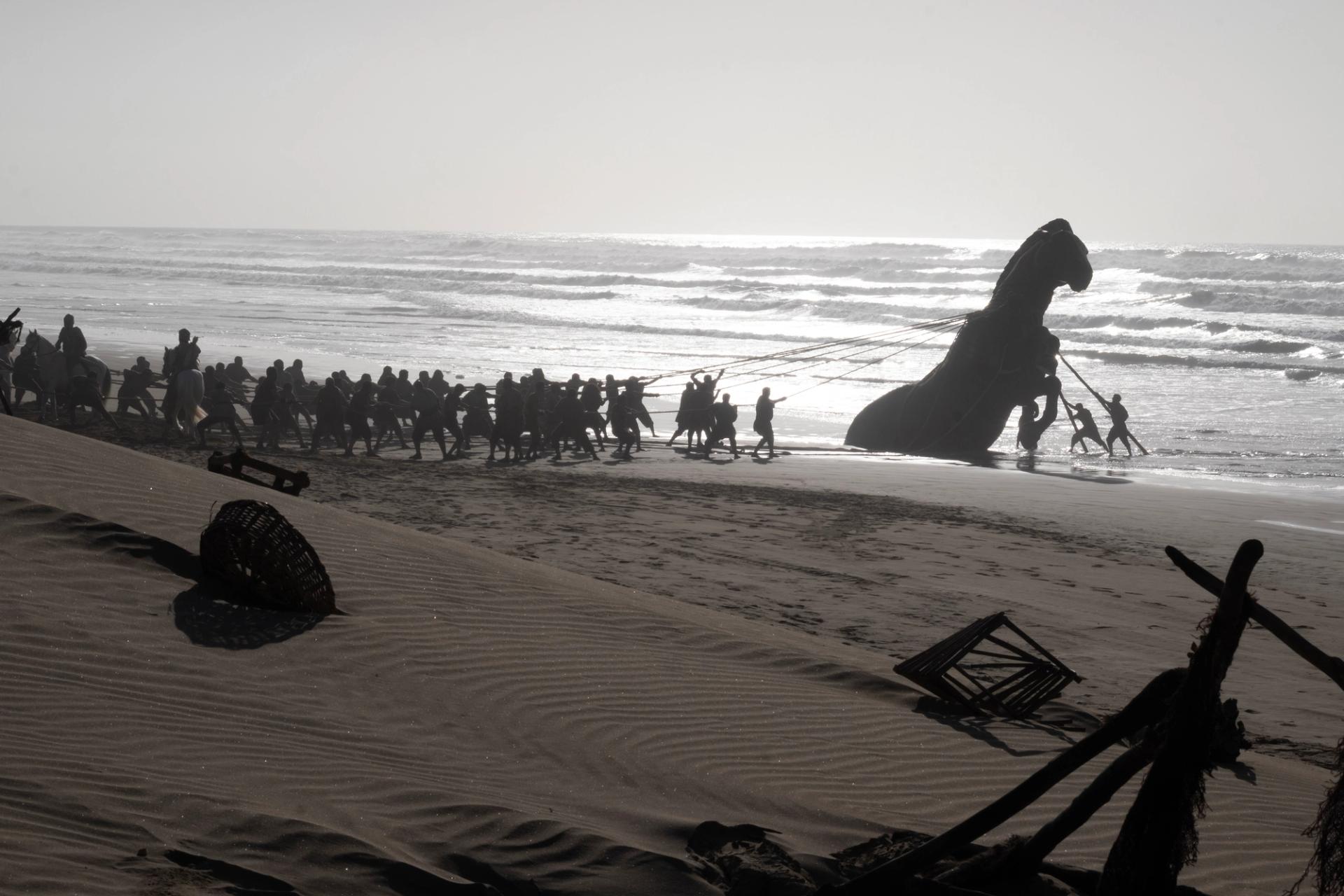 A black and white film still of a large crowd of figures hauling a massive horse sculpture from the ocean onto a beach using ropes.