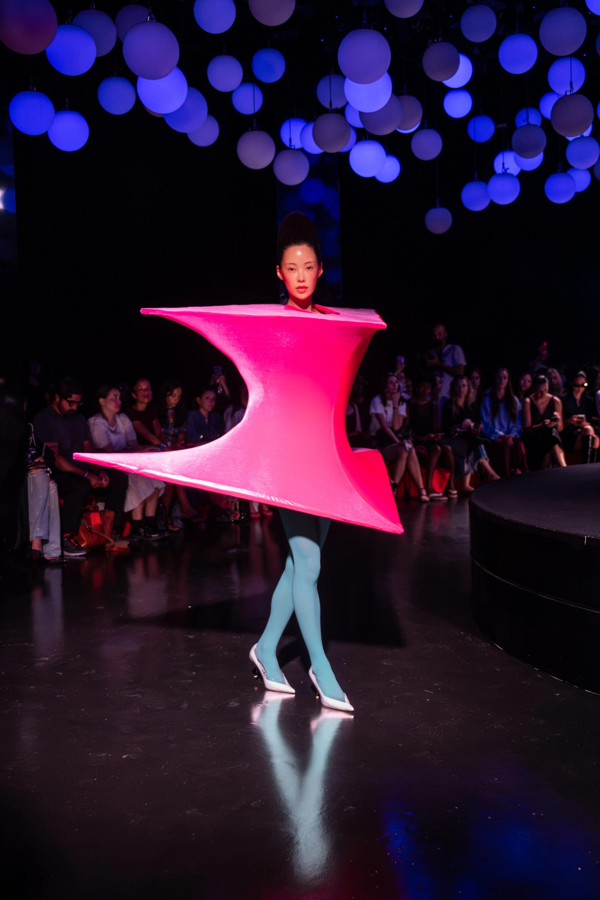 A model walks a darkened runway wearing a sculptural neon pink hourglass-shaped structured garment with teal tights and white heels, beneath a ceiling of glowing spheres