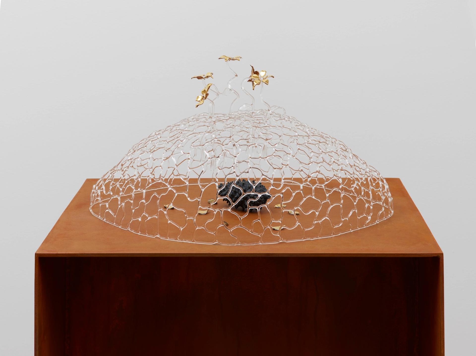 A dome-shaped glass latticework sculpture on a rust-colored pedestal, with small gold floral and bird forms emerging from its top and a dark mineral at its center.