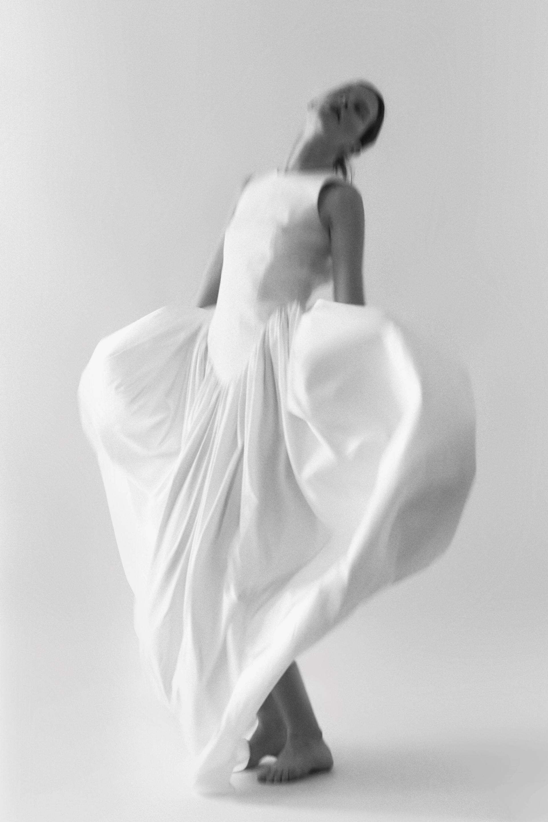 A blurry black-and-white image of a woman in floor-length white dress holding her skirt away from her body