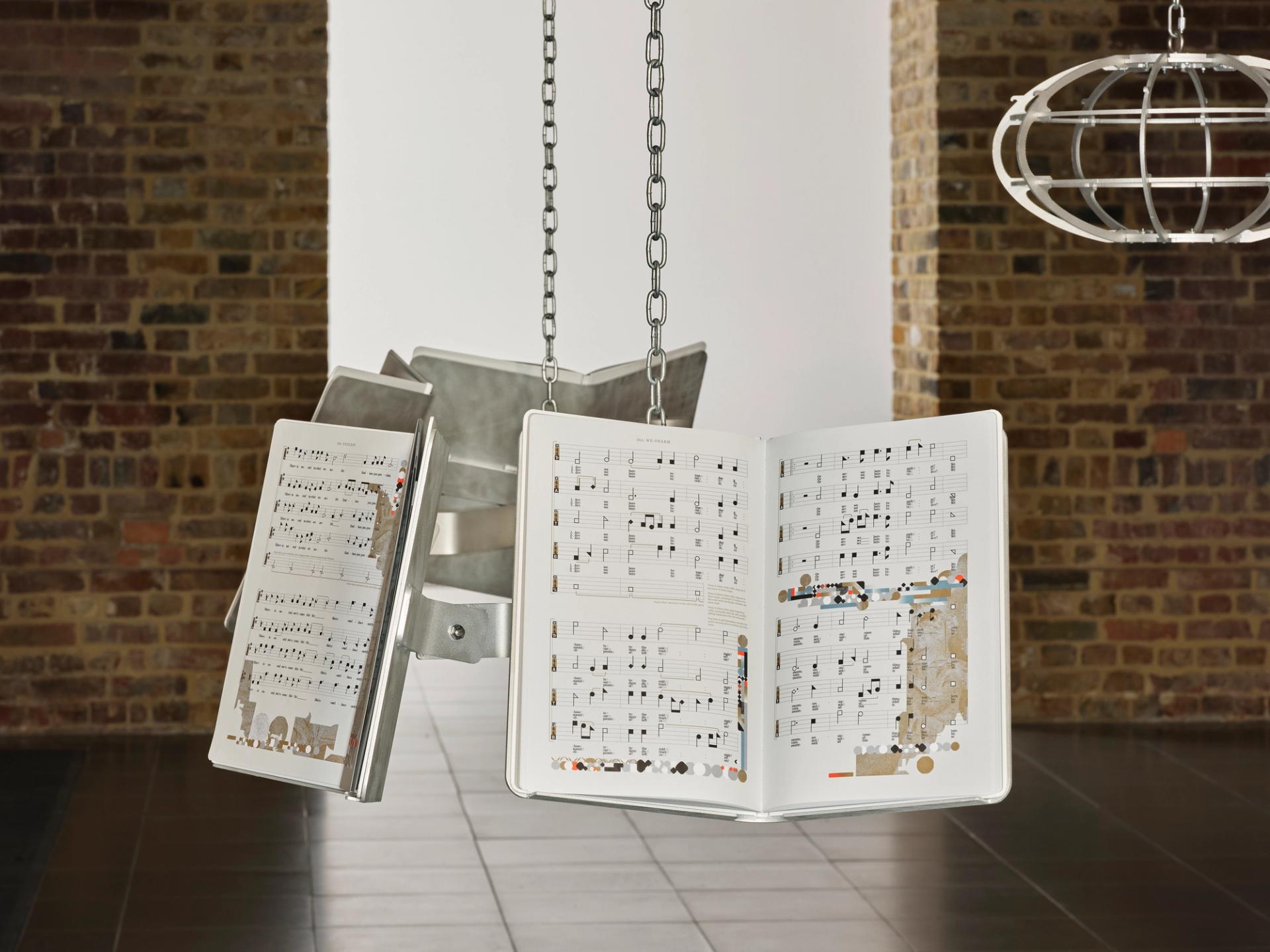 An art installation in a brick-walled gallery: oversized open books displaying collaged sheet music hang from metal chains attached to a sculptural metal form.