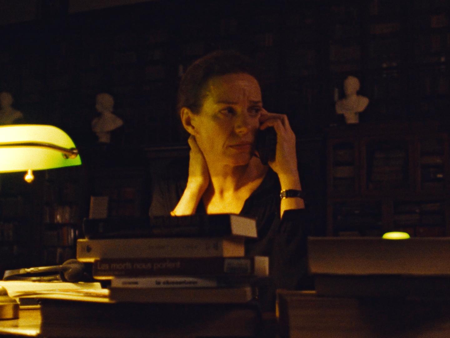 Dimly lit color film still of a woman on the phone in a wood-paneled library at night, lit by a green banker’s lamp