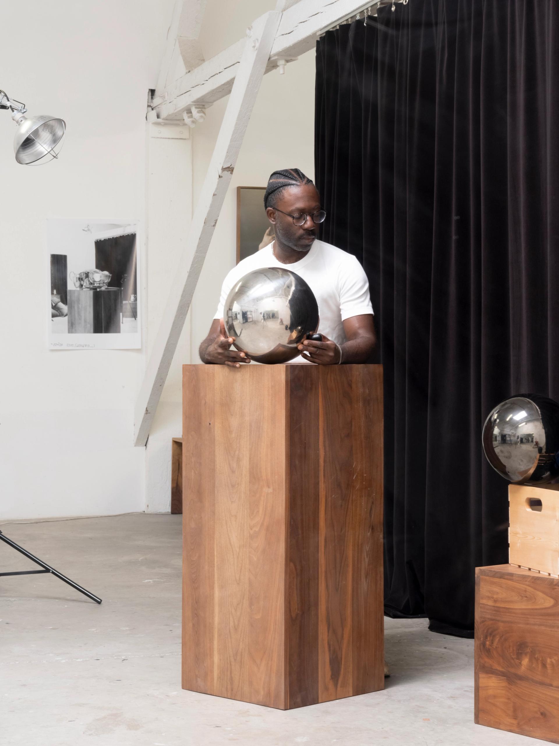 Person stands behind a wooden pedestal holding a reflective sphere in a studio setting.