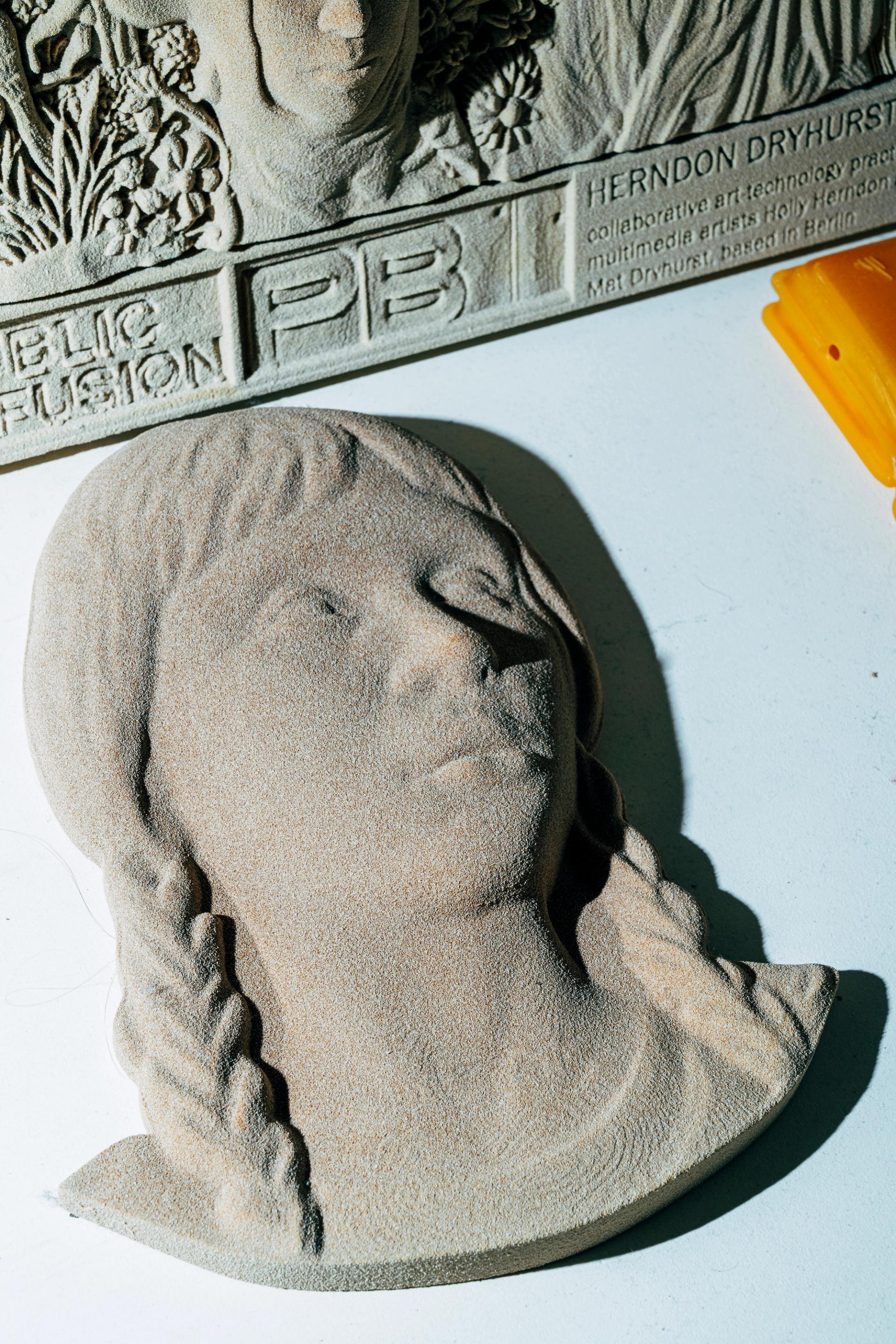 Two 3D-printed sand-textured relief sculptures of faces, with text reading 'Herndon Dryhurst' and 'Public Fusion' visible on the upper piece.