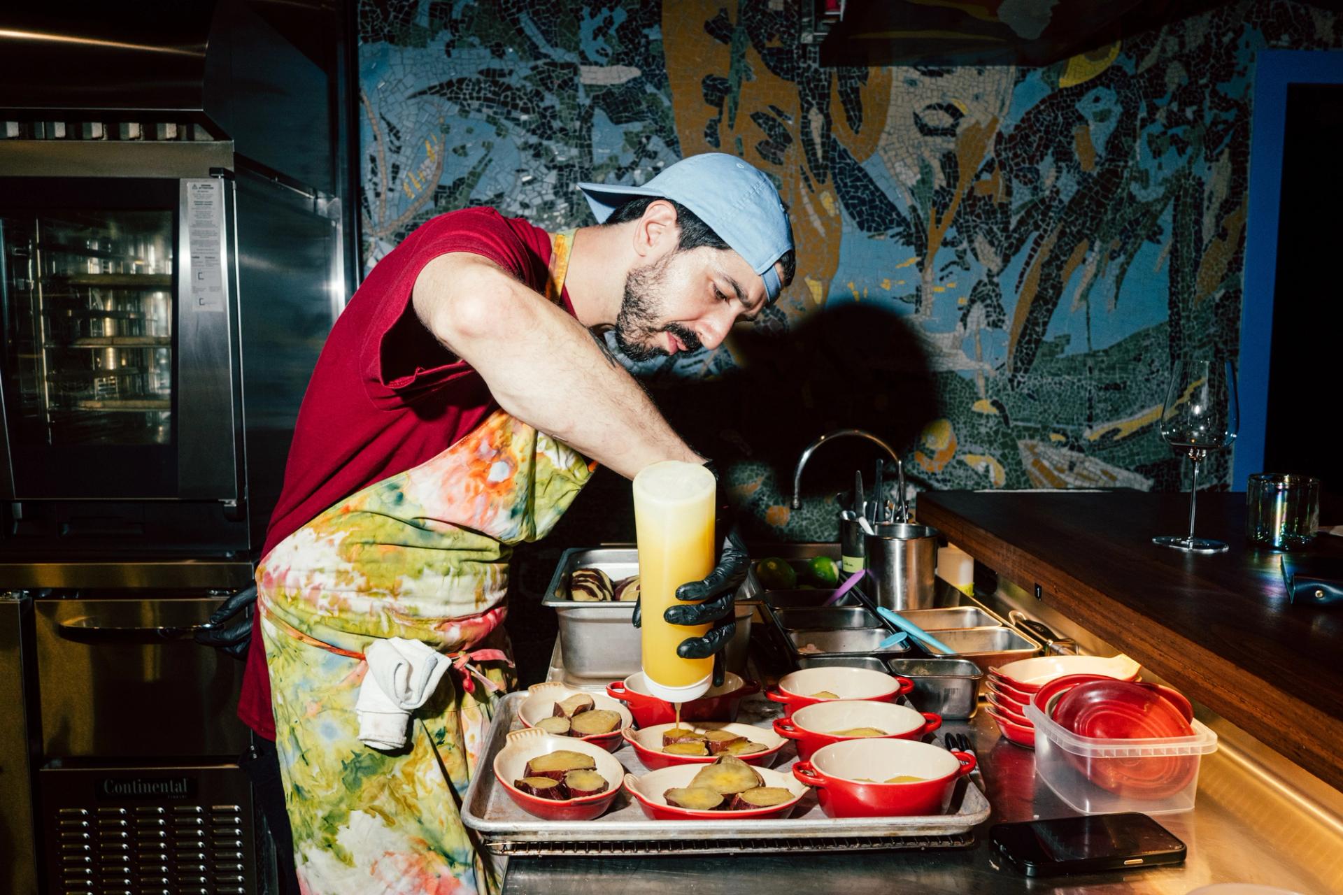 Man in a red shirt and blue hat pouring sauce from a squeeze bottle onto plated dishes.