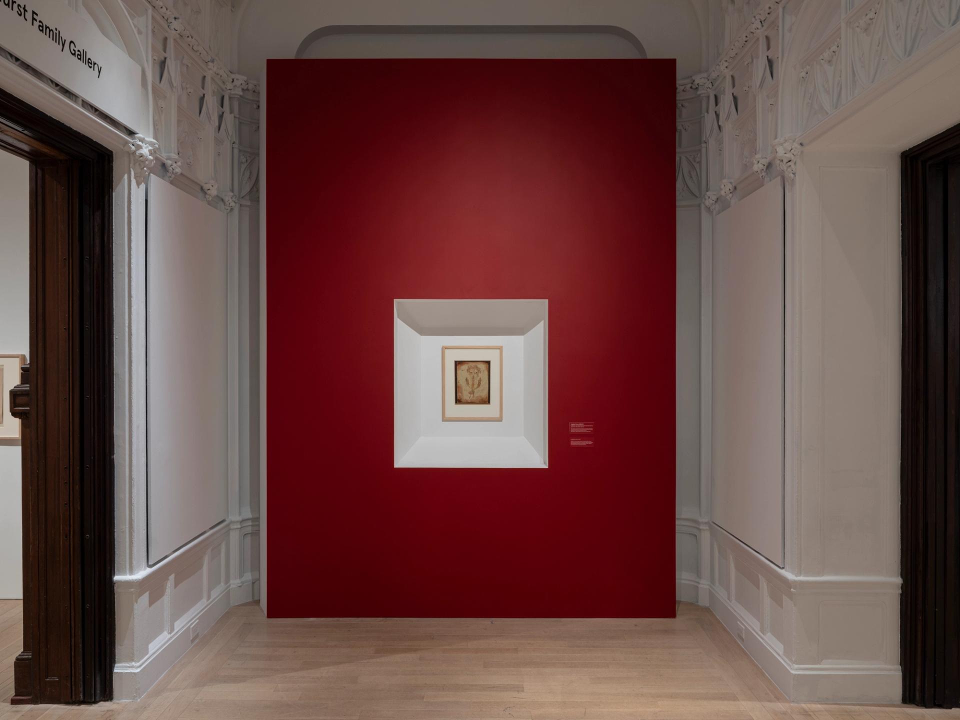A small framed artwork is displayed in a recessed niche within a large deep-red wall inside an ornate gallery space.