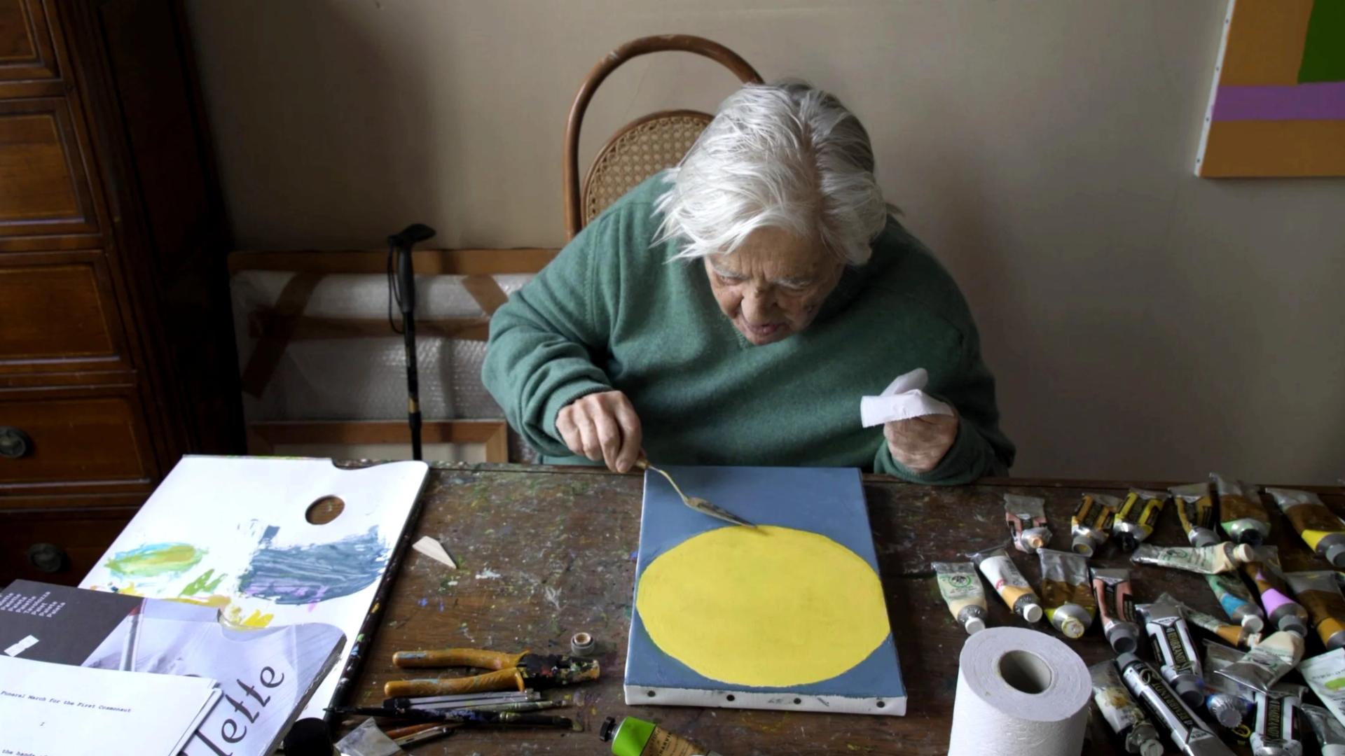 An elderly woman with white hair leans over a worktable, painting a yellow circle on a blue canvas, surrounded by paint tubes, brushes, and a palette.