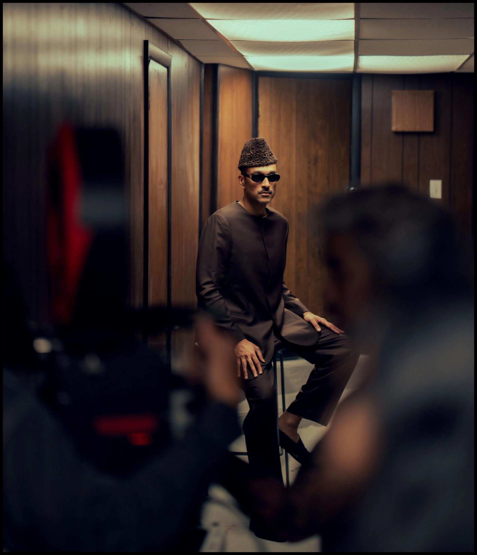 A behind-the-scenes film set photograph showing a man in a dark kurta, hat, and sunglasses seated in a wood-paneled room, with a camera and crew member visible in the foreground.