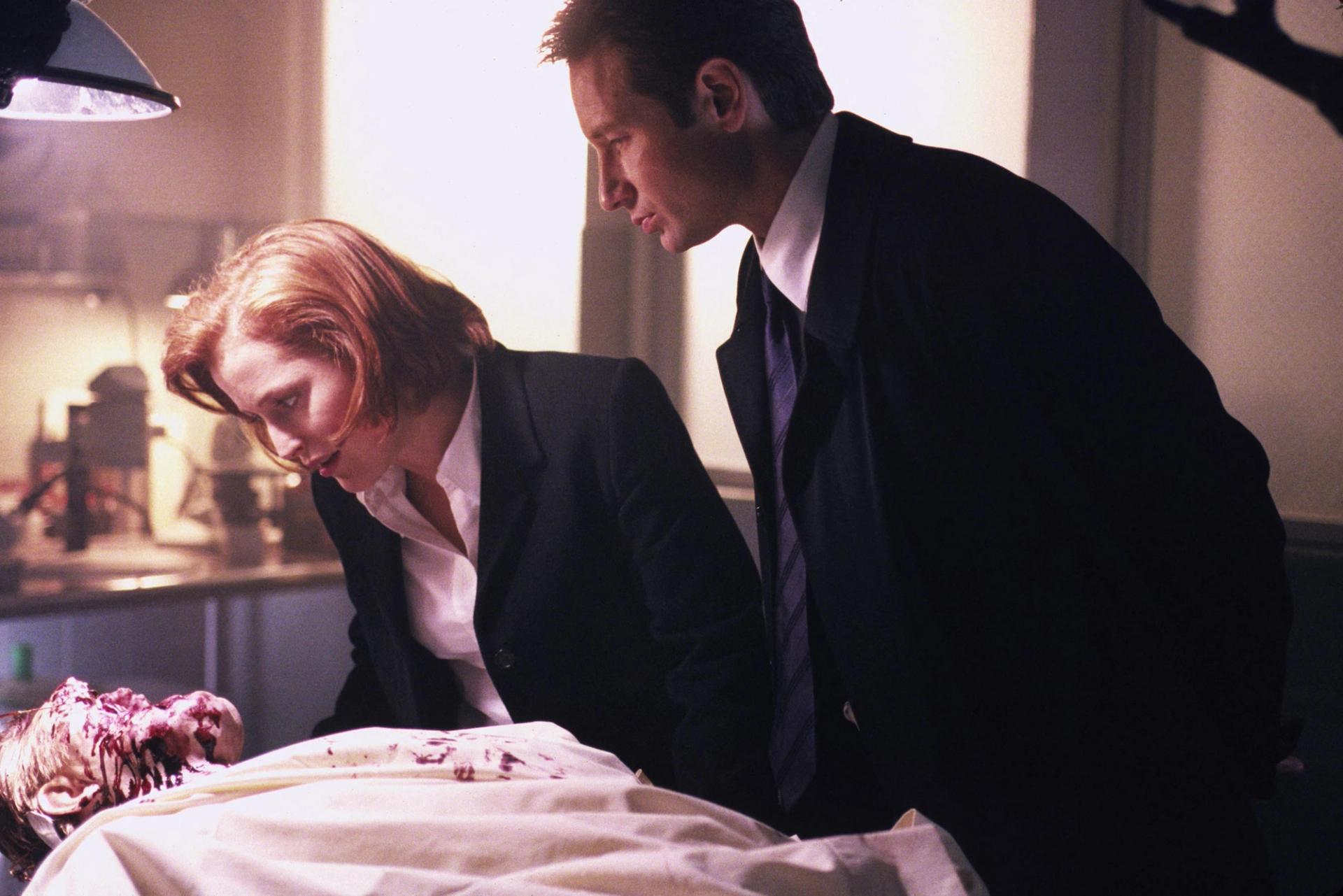 A man and woman in dark suits leaning over an examination table, looking down at a body with visible head wounds and blood on the sheet, in a dimly lit room.