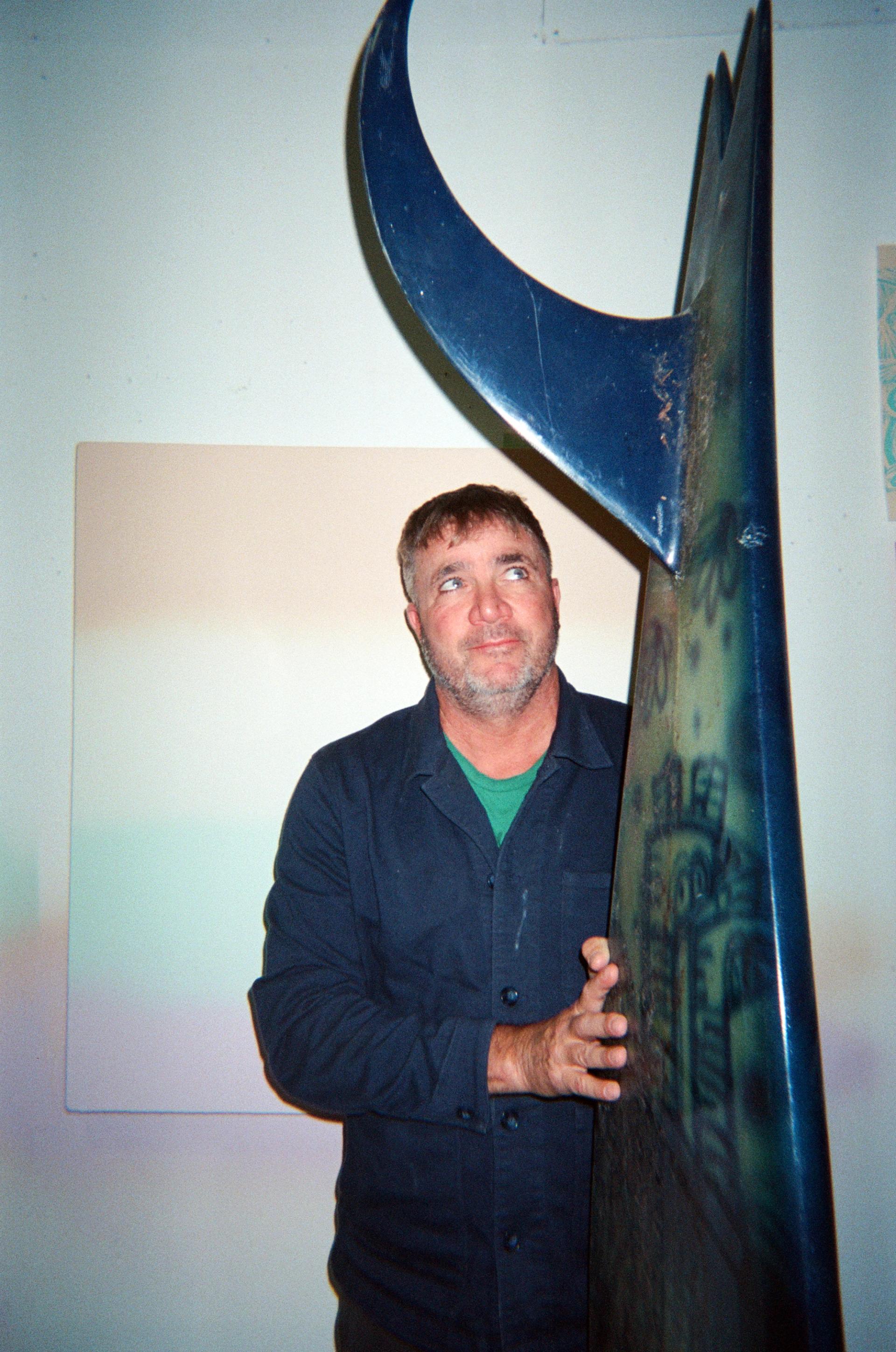 A bearded man in a navy work jacket holding up a surf board with a crescent-shaped fin, with a pastel-striped painting visible on the wall behind him