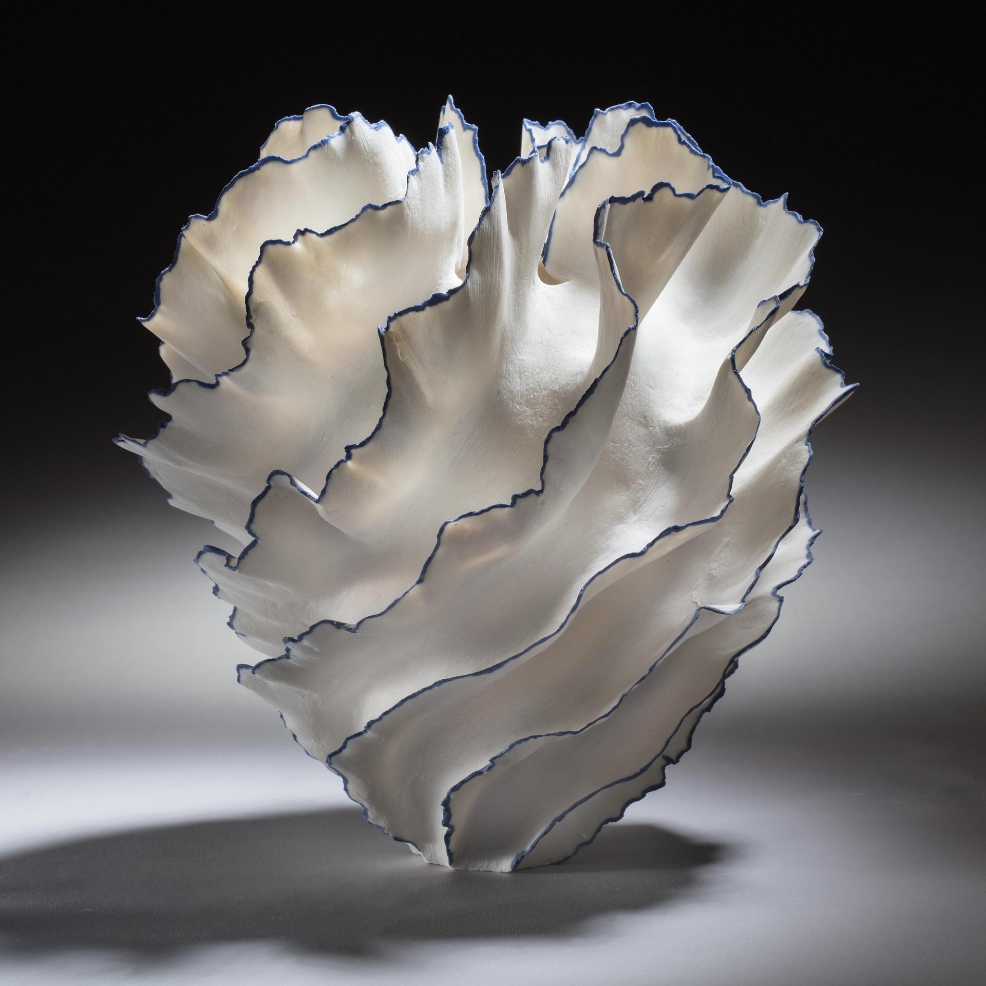 White ruffled ceramic vessel with blue-edged, petal-like layers, photographed against a dark background.