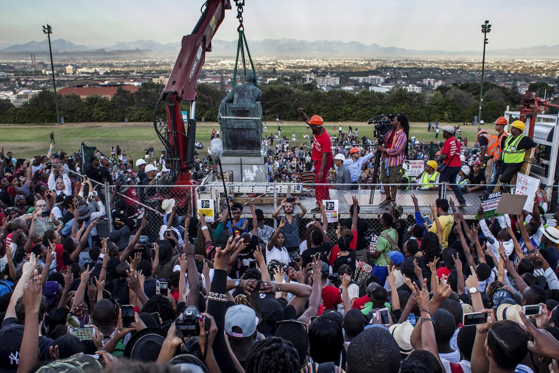 A large crowd with raised hands cheers as a bronze statue is lifted by a crane, surrounded by construction workers and protesters, with a city and mountains visible in the background.