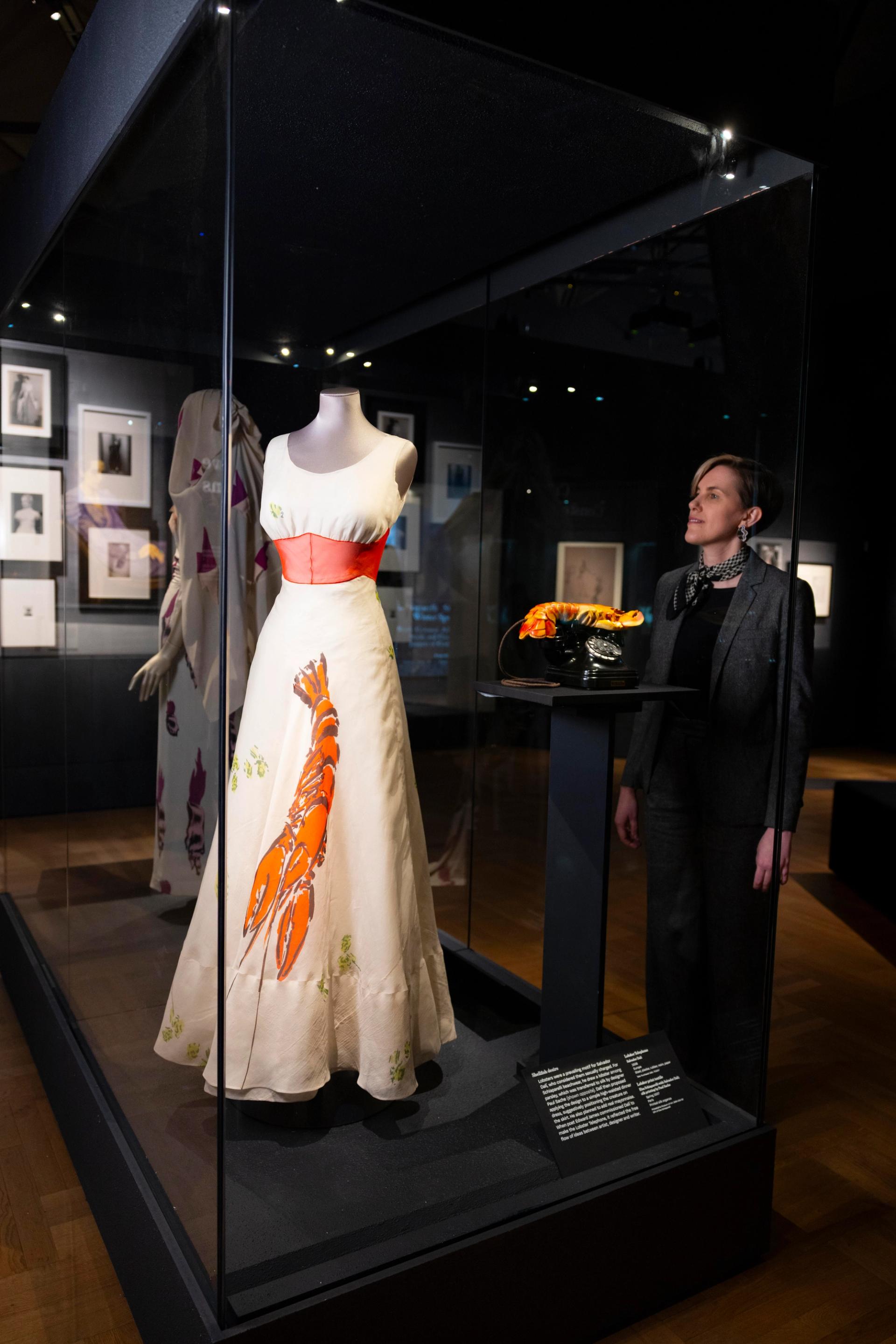 Schiaparelli lobster-print gown displayed with Salvador Dalí's "Lobster Telephone" in a glass case at the V&A museum, viewed by a visitor.