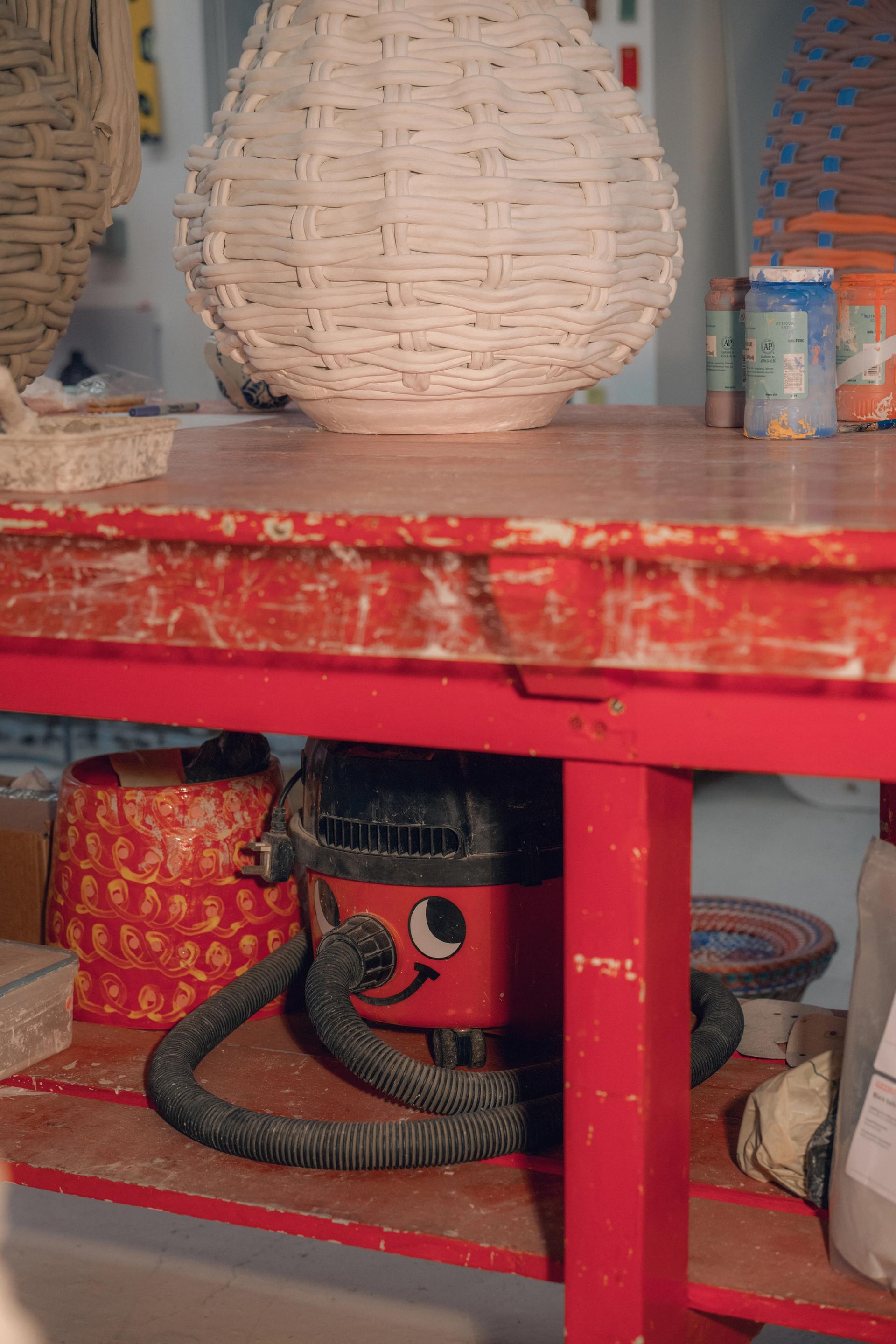 A red wooden table with an unfinished woven clay sculpture on top and a vacuum with sticker eyes underneath.