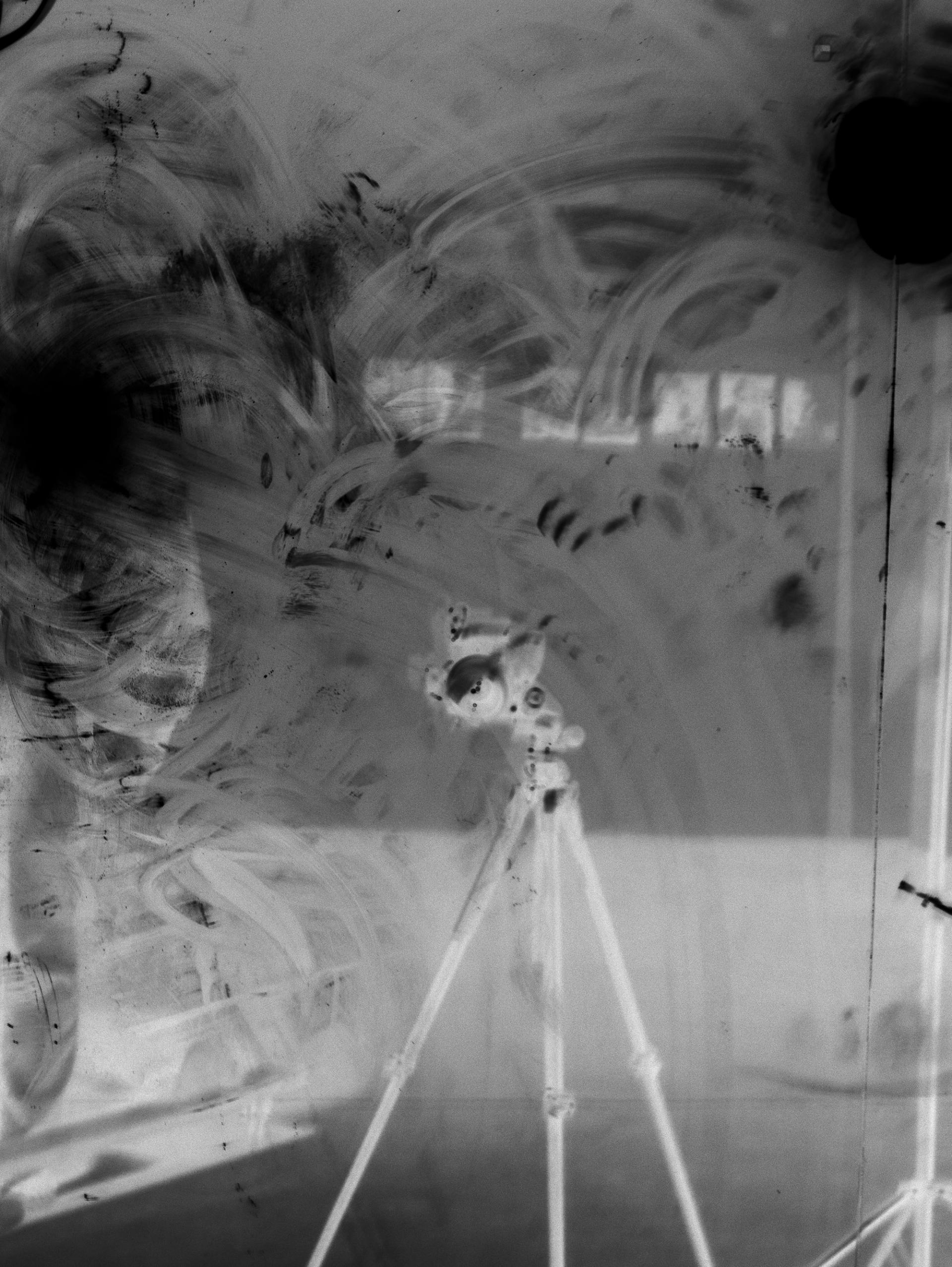 Black-and-white negative image of a smudged mirror reflecting a tripod-mounted camera.