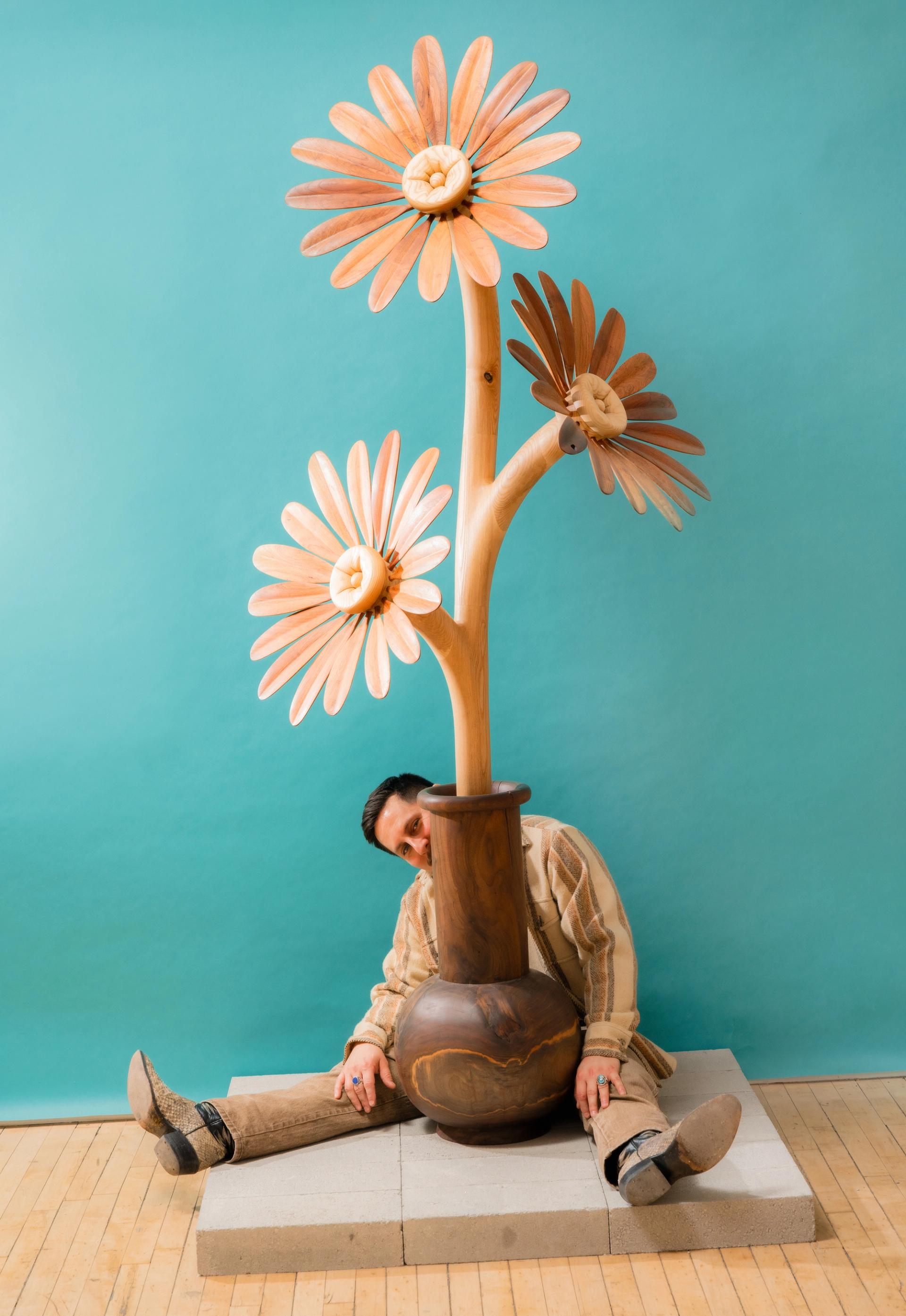 A man seated on the floor peeks out from behind a large sculpture of a potted flower