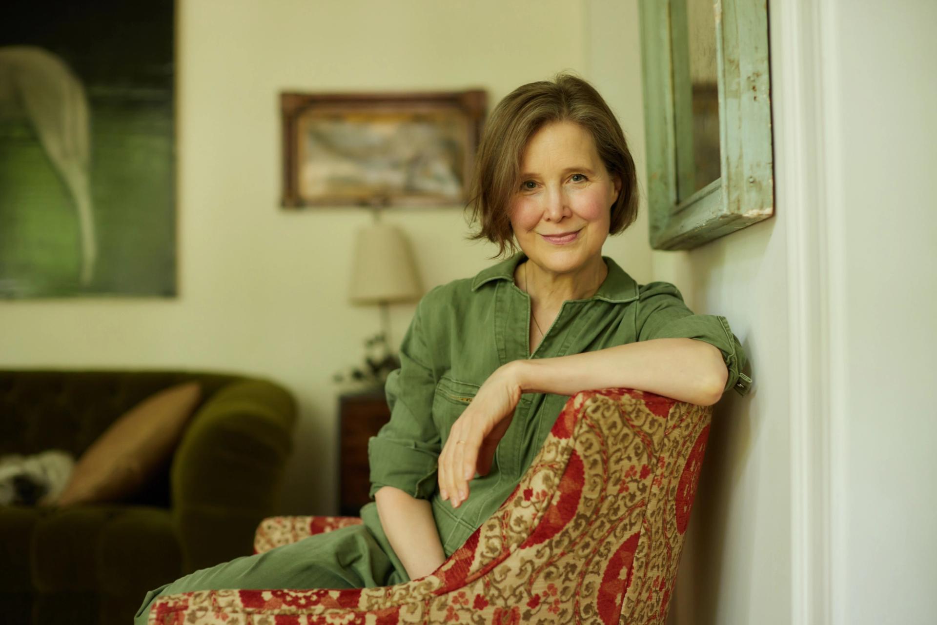 A woman in a green jumpsuit sits in a patterned armchair, smiling gently in a cozy living room.