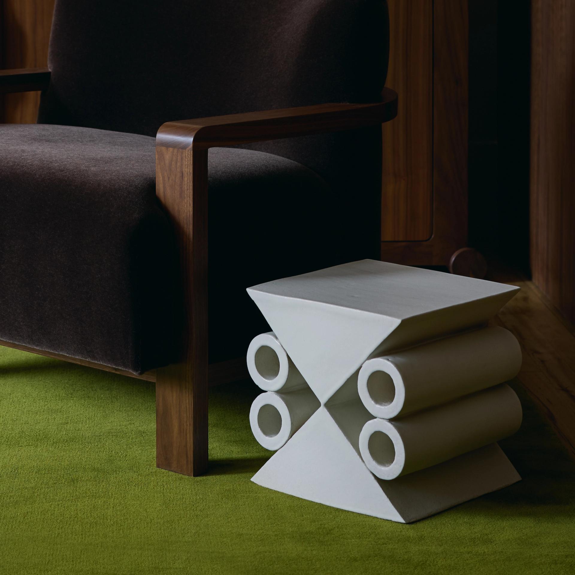 White geometric ceramic side table beside upholstered armchair on green carpeted floor.