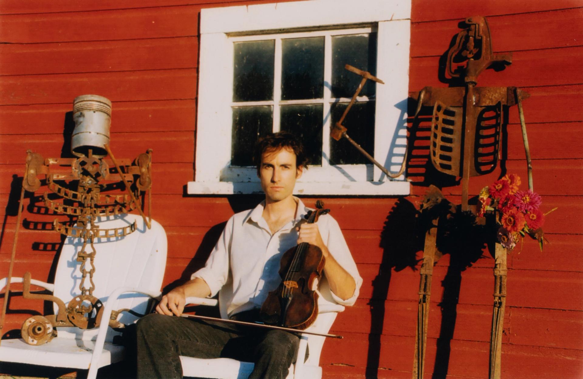 A white man with brown hair sits holding a violin between two metal skeleton sculptures.