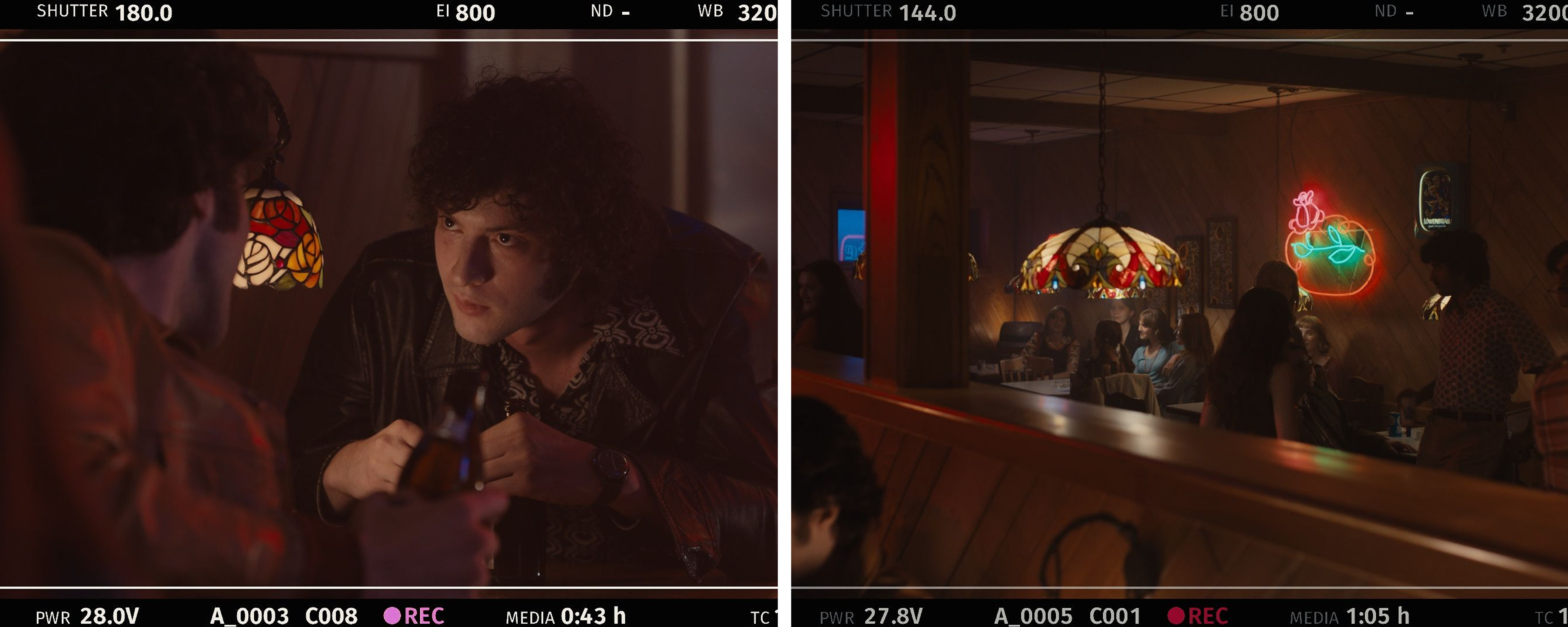 Behind-the-scenes still from a camera monitor showing a curly-haired man in a leather jacket holding a beer bottle across a table from another man seen from behind, lit by a stained-glass lamp; Behind-the-scenes still from a camera monitor showing a dimly lit, retro-style bar scene: patrons seated at wood-paneled booths under a stained-glass pendant lamp, with neon beer and rose signs glowing on the wall