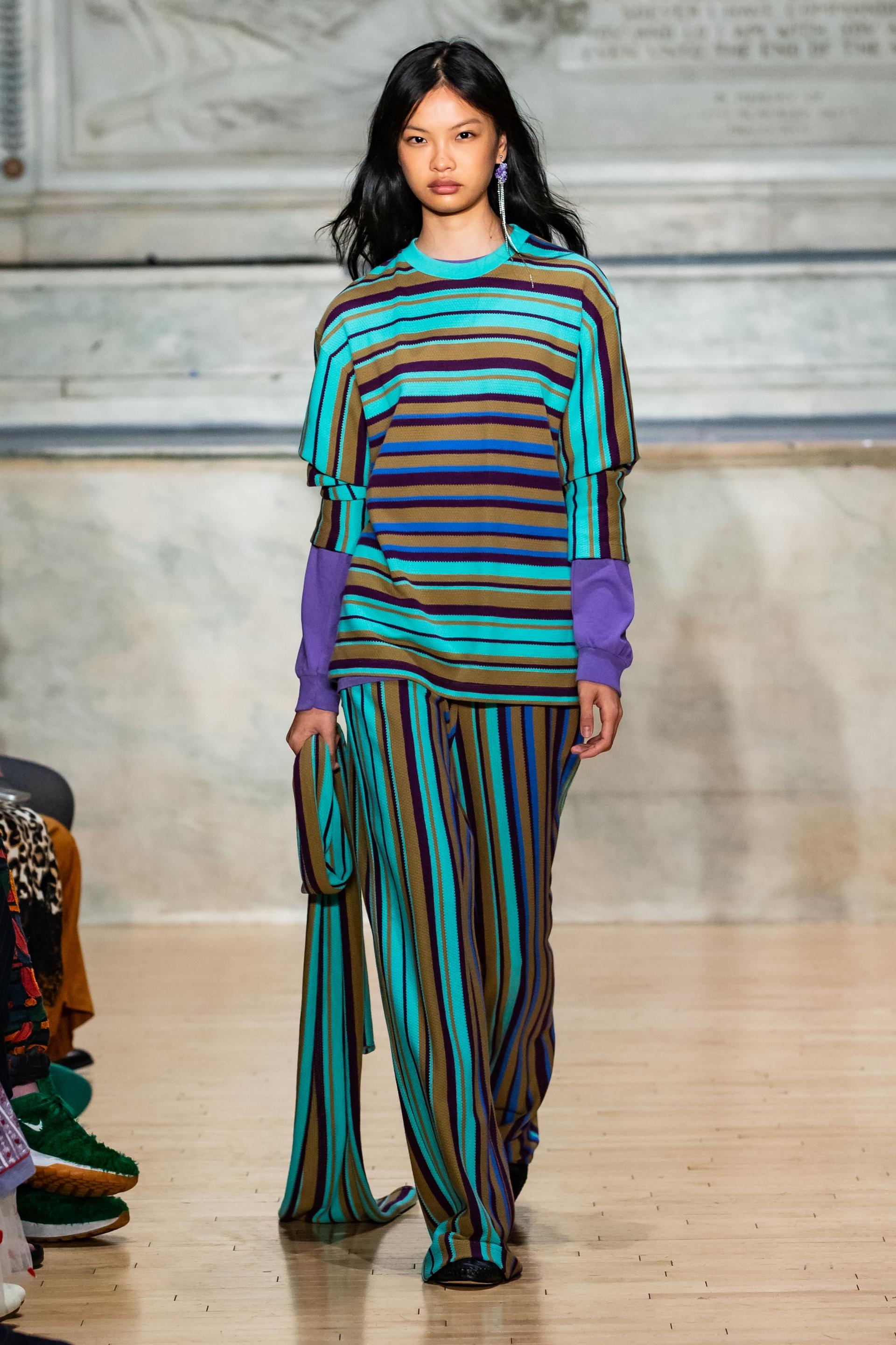 A model walks a runway in a bold multicolored striped knit set — a horizontal-striped top layered over a long-sleeve purple shirt, paired with vertical-striped wide-leg trousers in matching tones