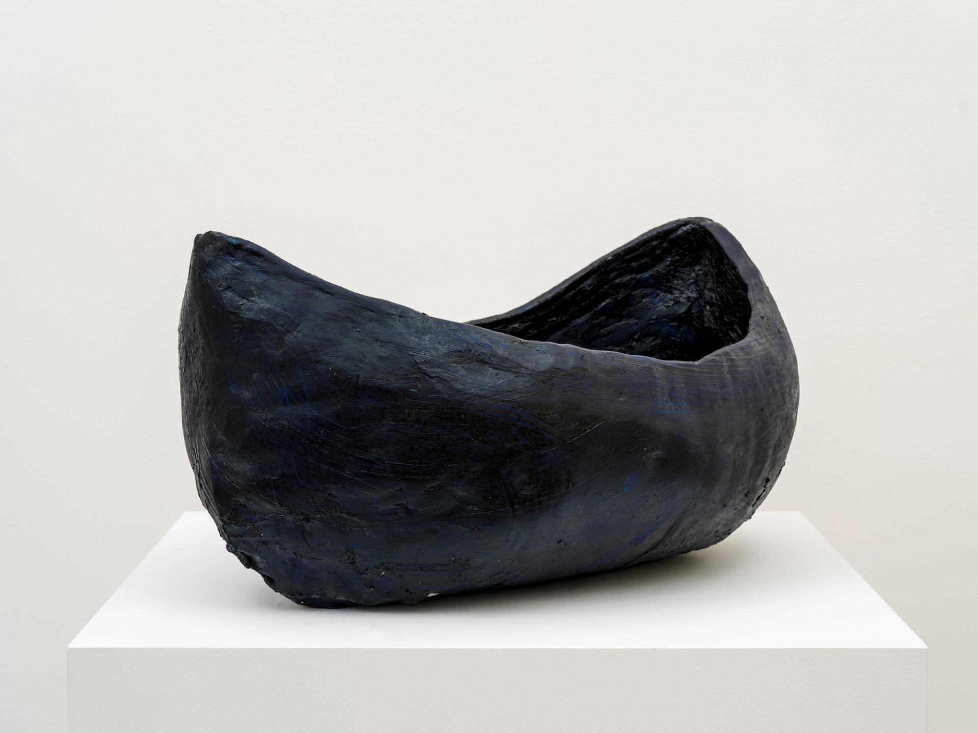 Dark, boat-shaped sculptural vessel displayed on white pedestal against gallery wall.
