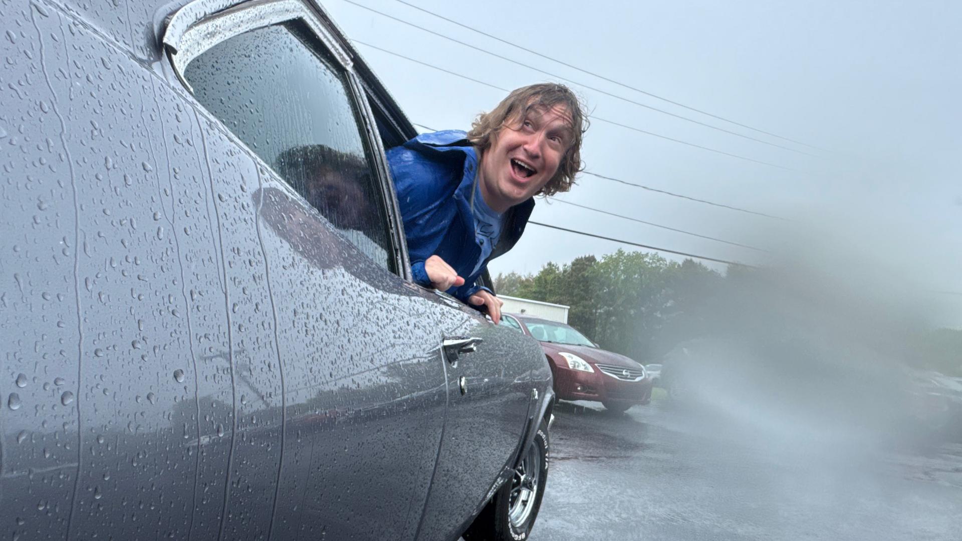 A laughing man with windblown hair leaning out of a vintage car’s window in the rain